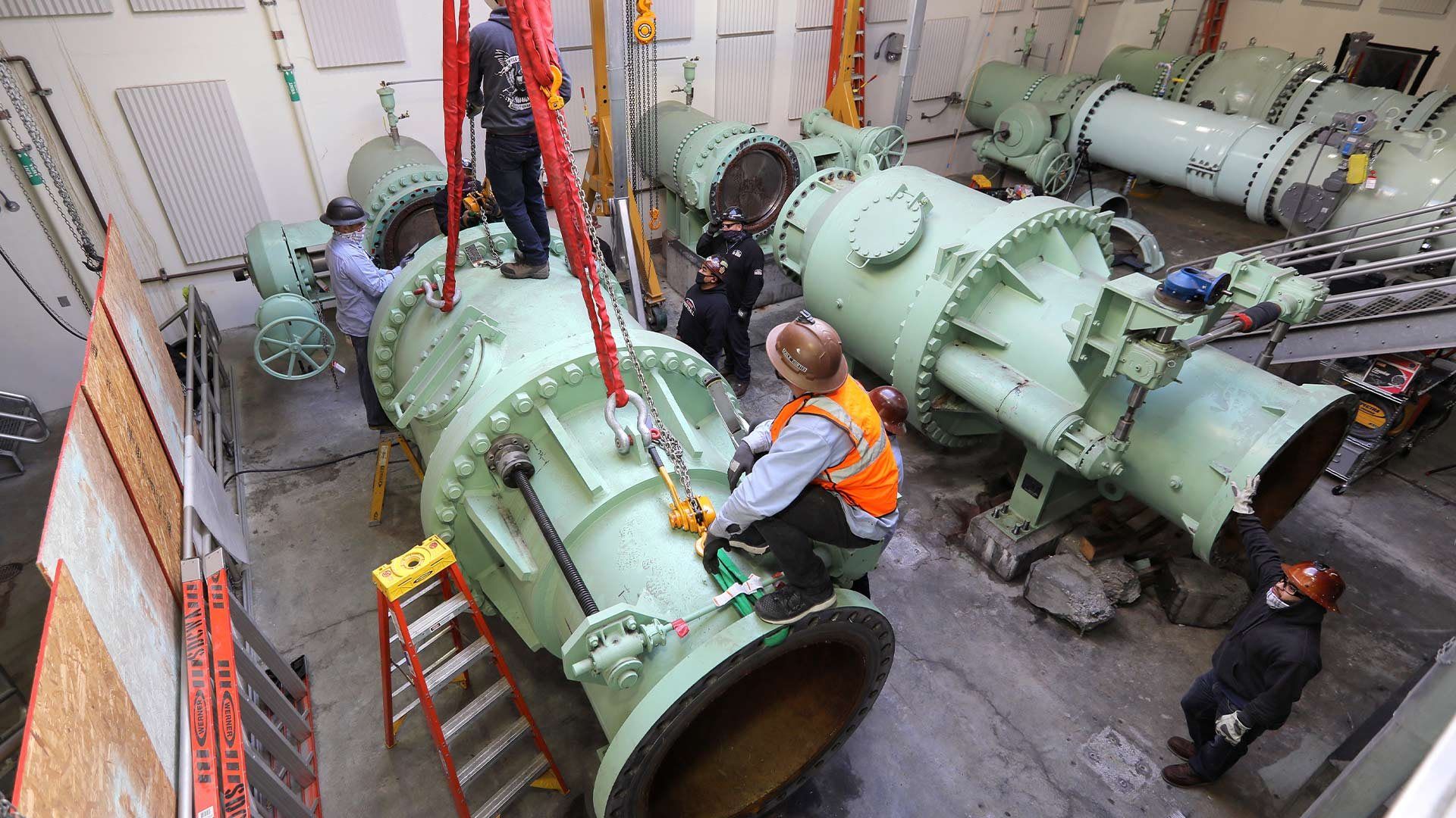 Workers in safety gear install or maintain large industrial water valves inside a facility, using heavy-duty lifting equipment and tools.