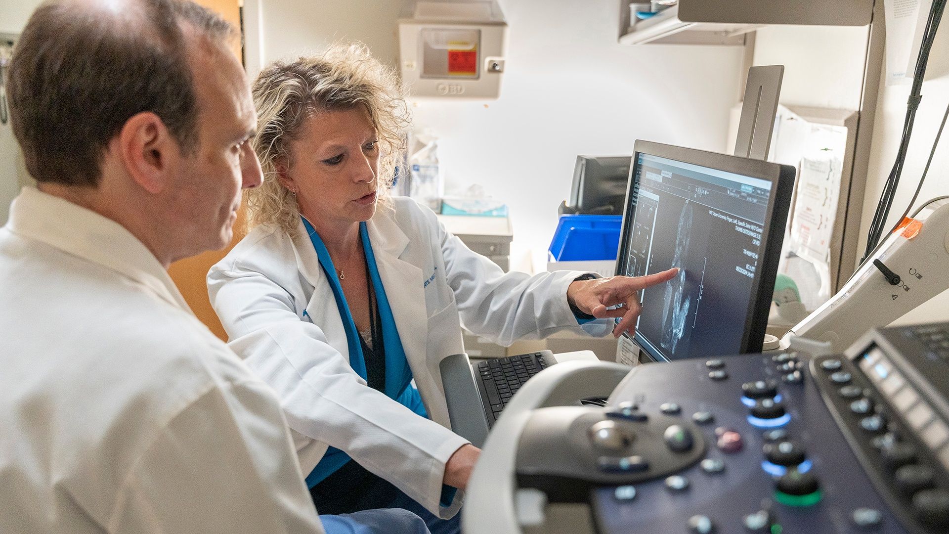 Two medical professionals in white coats reviewing a scan on a computer monitor in a clinical setting, with one pointing at the image displayed on the screen.