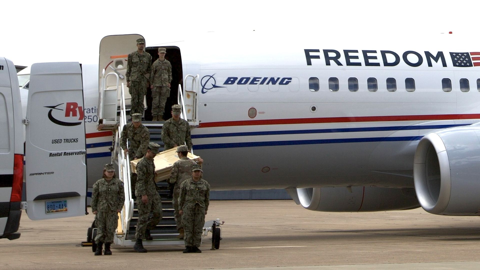 Several soldiers in camouflage carry a coffin down mobile stairs from a Boeing cargo plane marked "FREEDOM" with an American flag on the side, parked on a tarmac.