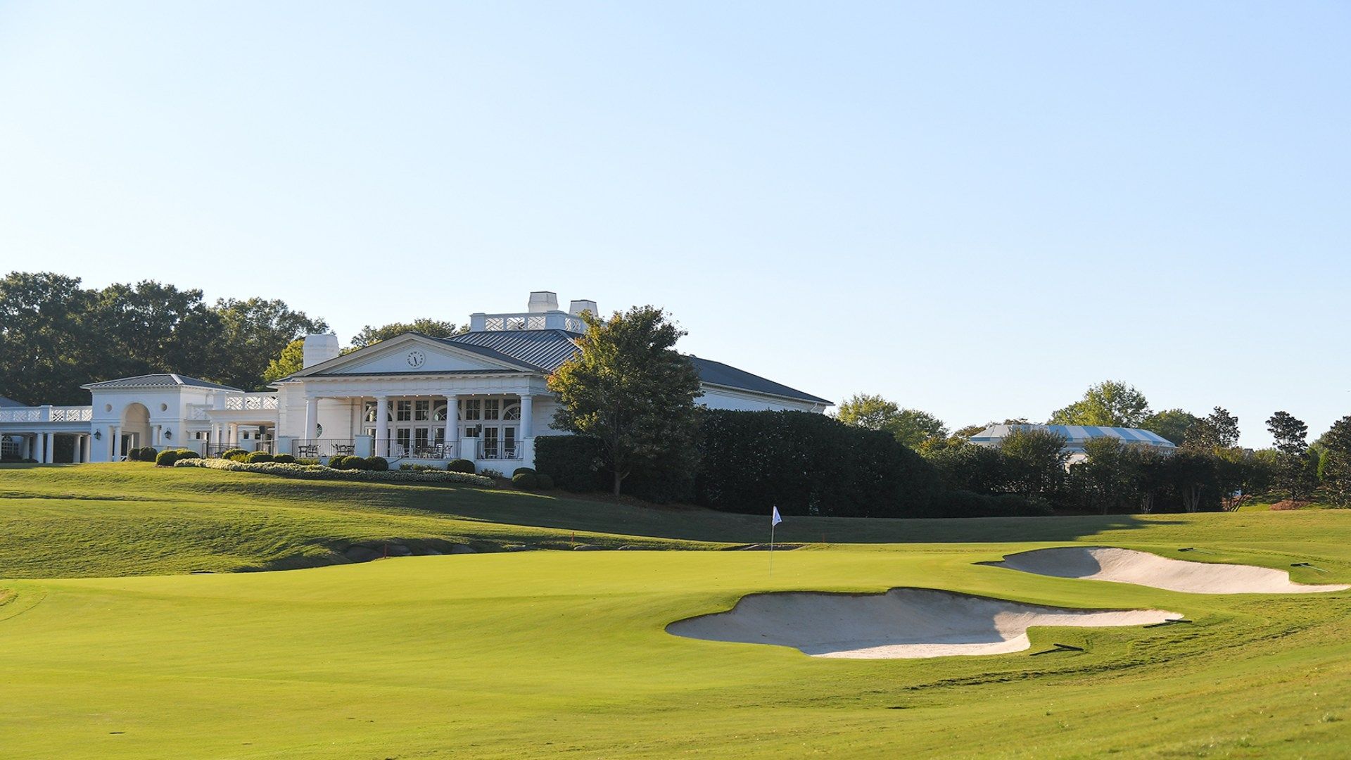 A white clubhouse sits beside a manicured golf course with green fairways and sand bunkers, under a clear blue sky.