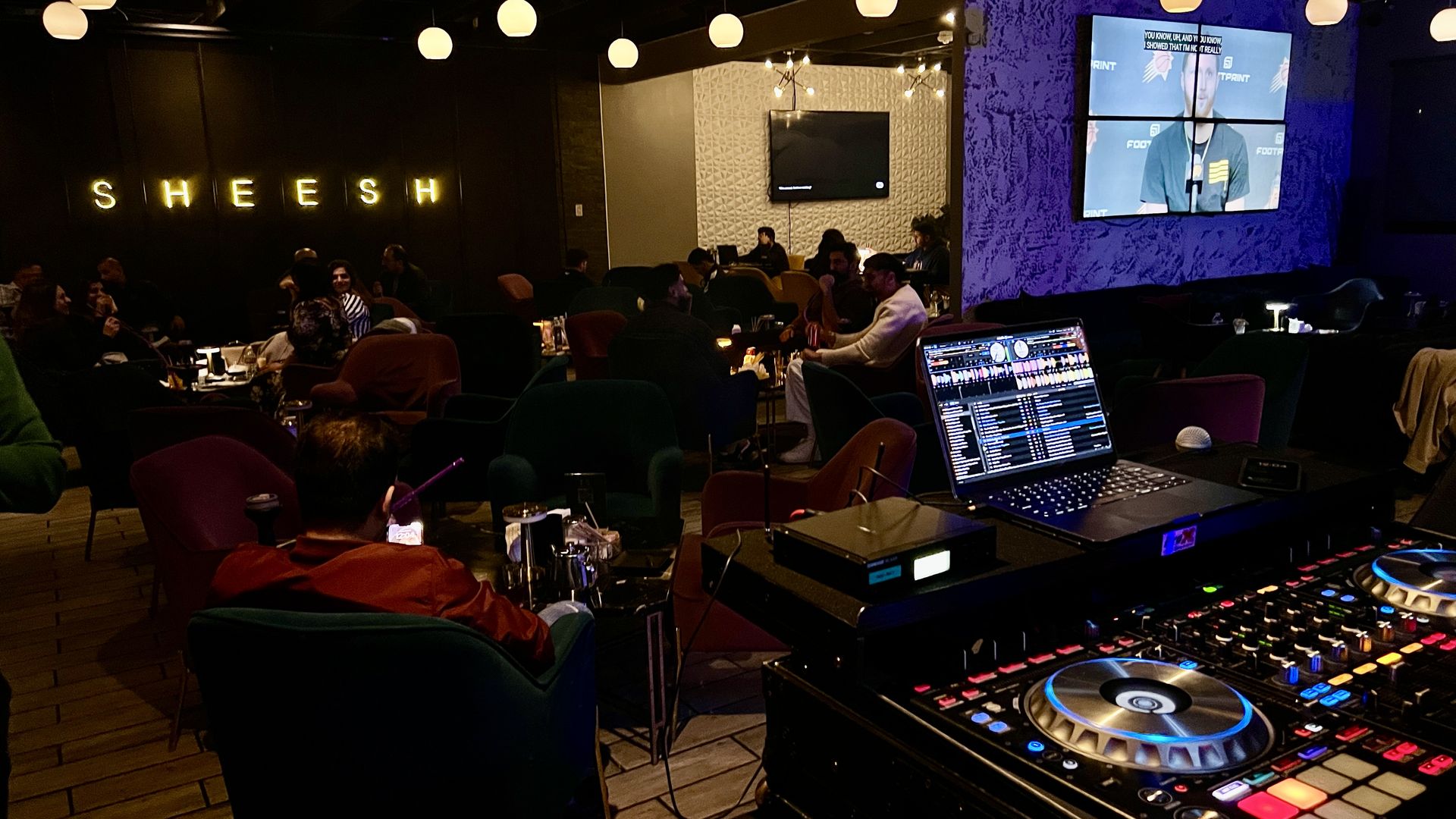 A lounge with a DJ booth and SHEESH written in neon lighting