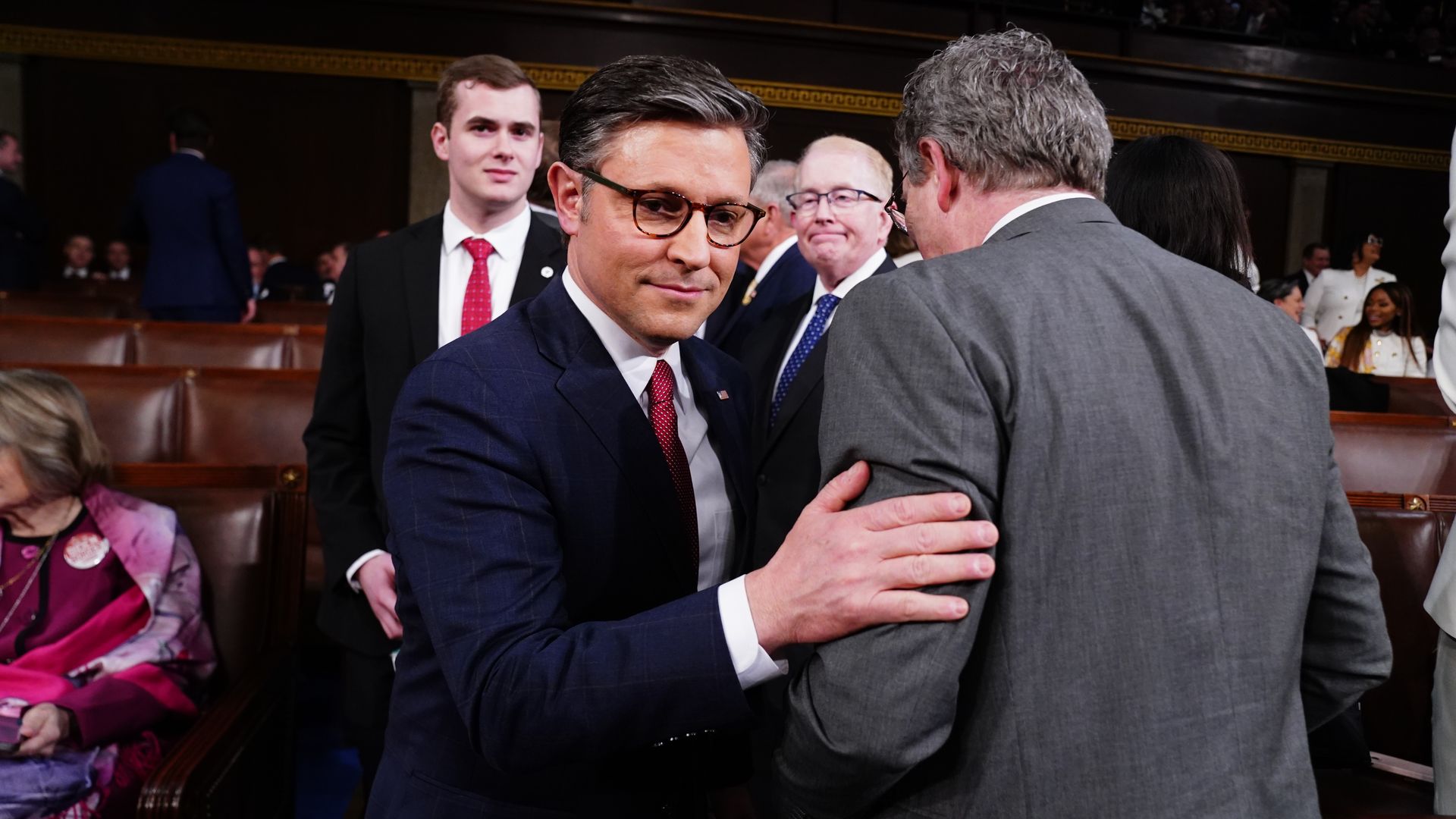 House Speaker Mike Johnson, wearing a blue suit, with his hand on the arm of Rep. Thomas Massie inside the House chamber.
