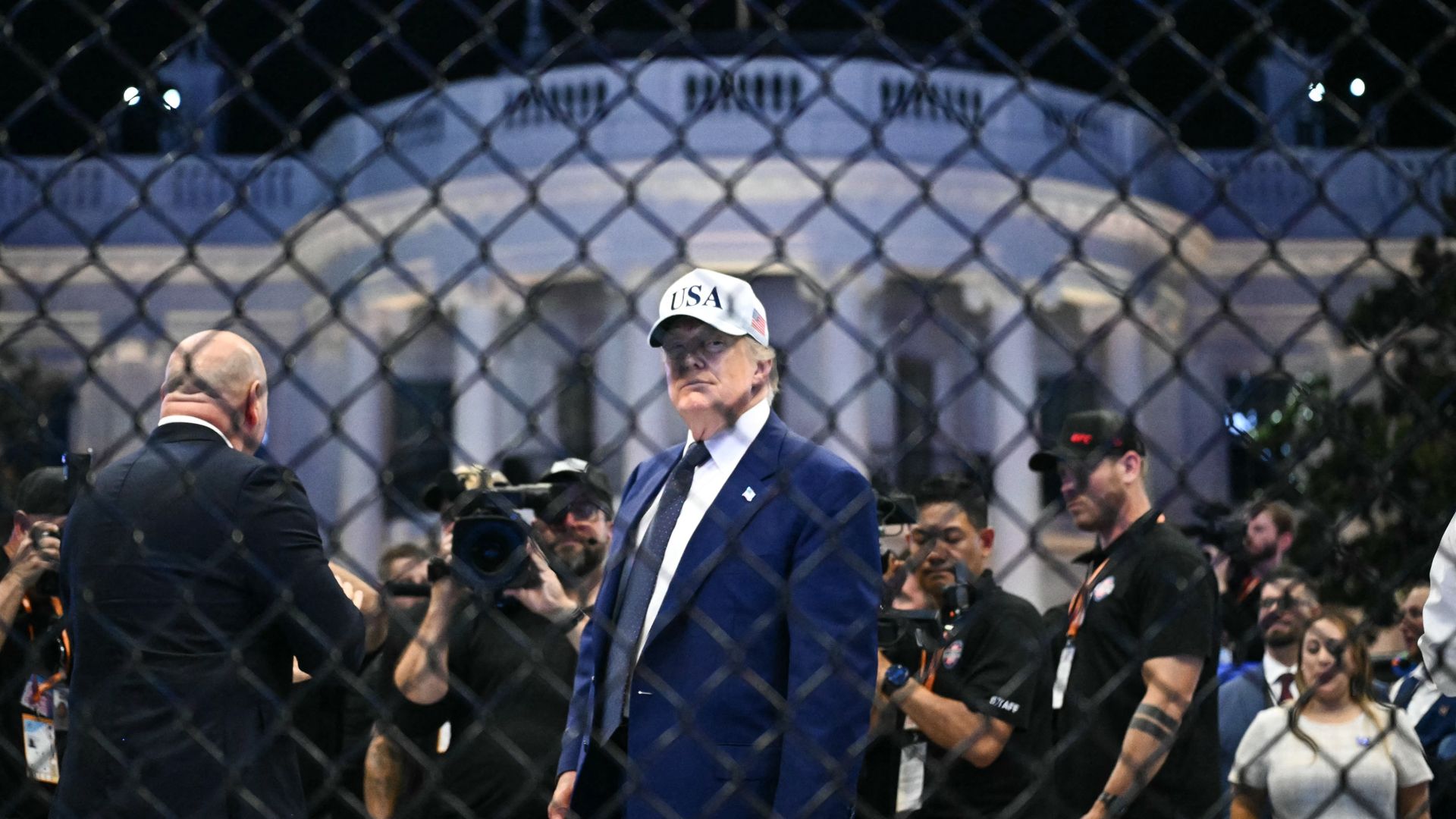 President Trump stands behind a fence during his UFC event at the White House, wearing a dark blue suit, a white collared shirt, a dark tie, an American flag pin and a white hat that says "USA" on it.