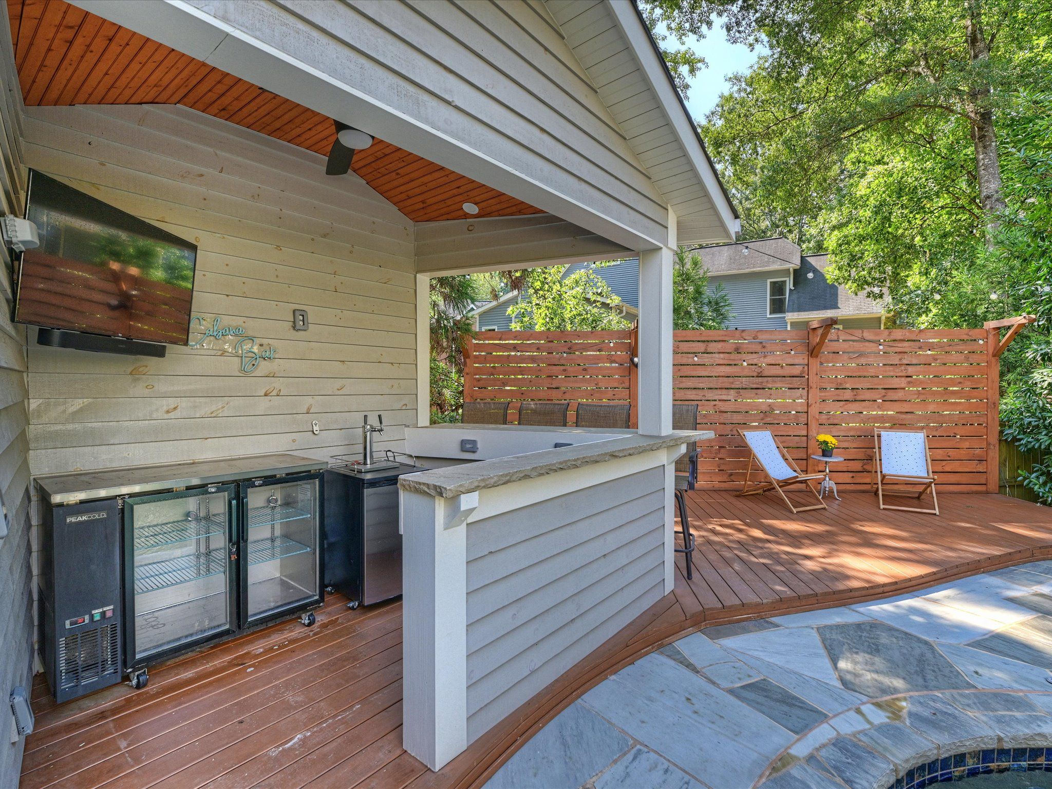 Outdoor patio with a wooden cabana bar, two glass-door refrigerators, a mounted TV, wooden deck, two white lounge chairs, a small table with yellow flowers, and a stained glass pool edge.
