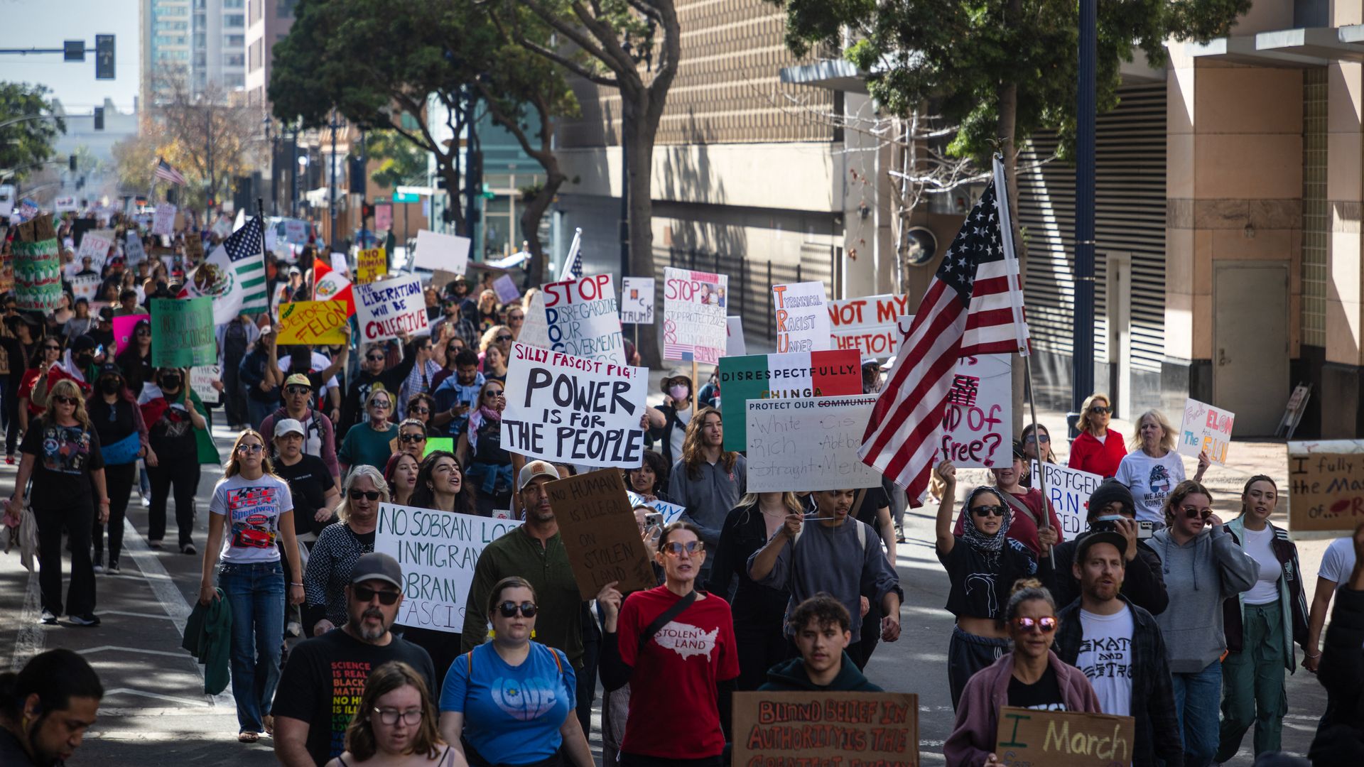 Demonstrators holding signs and flags march through the street in protest of President Donald Trump's policies.