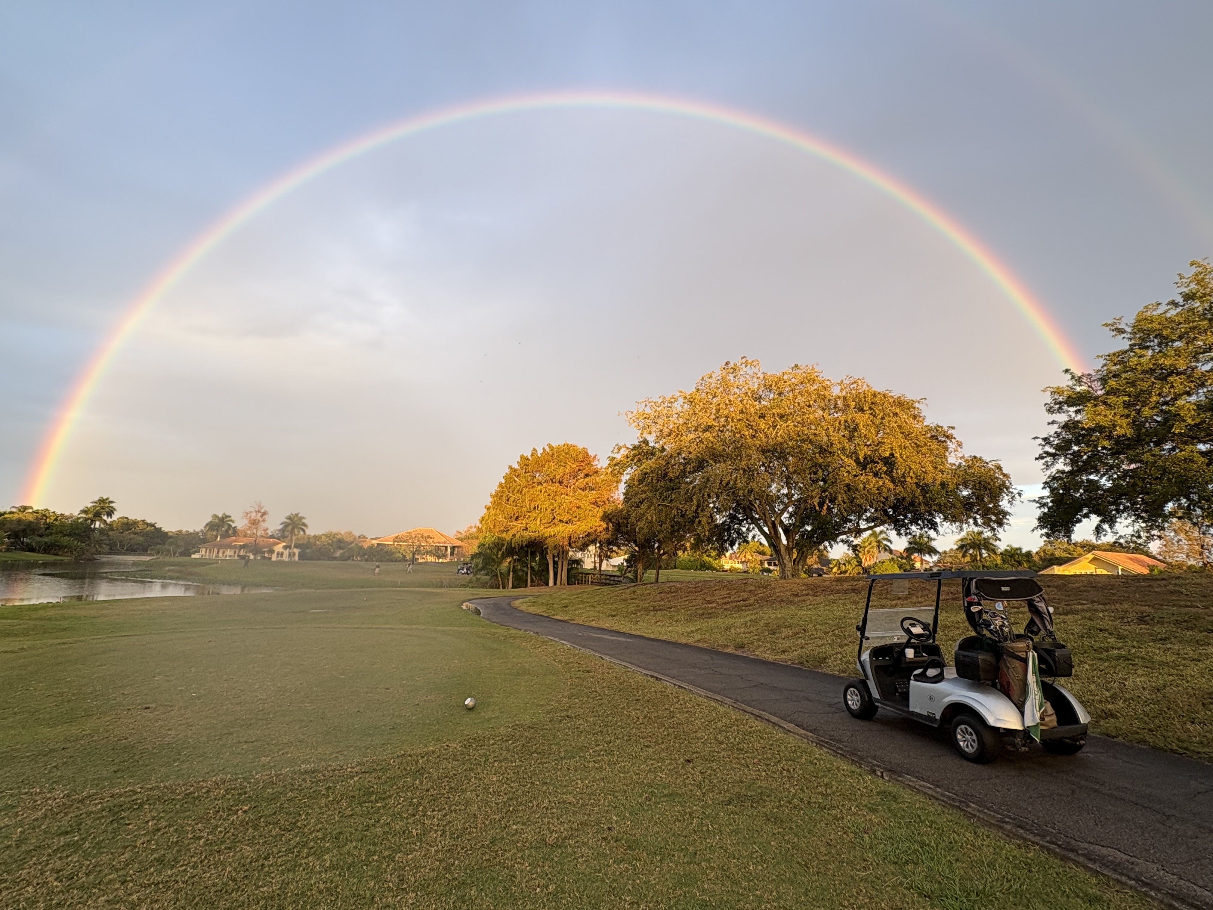 Golf course with a silver golf cart on a paved path under a bright, full rainbow arching across a partly cloudy sky during golden hour with trees and houses in the background.