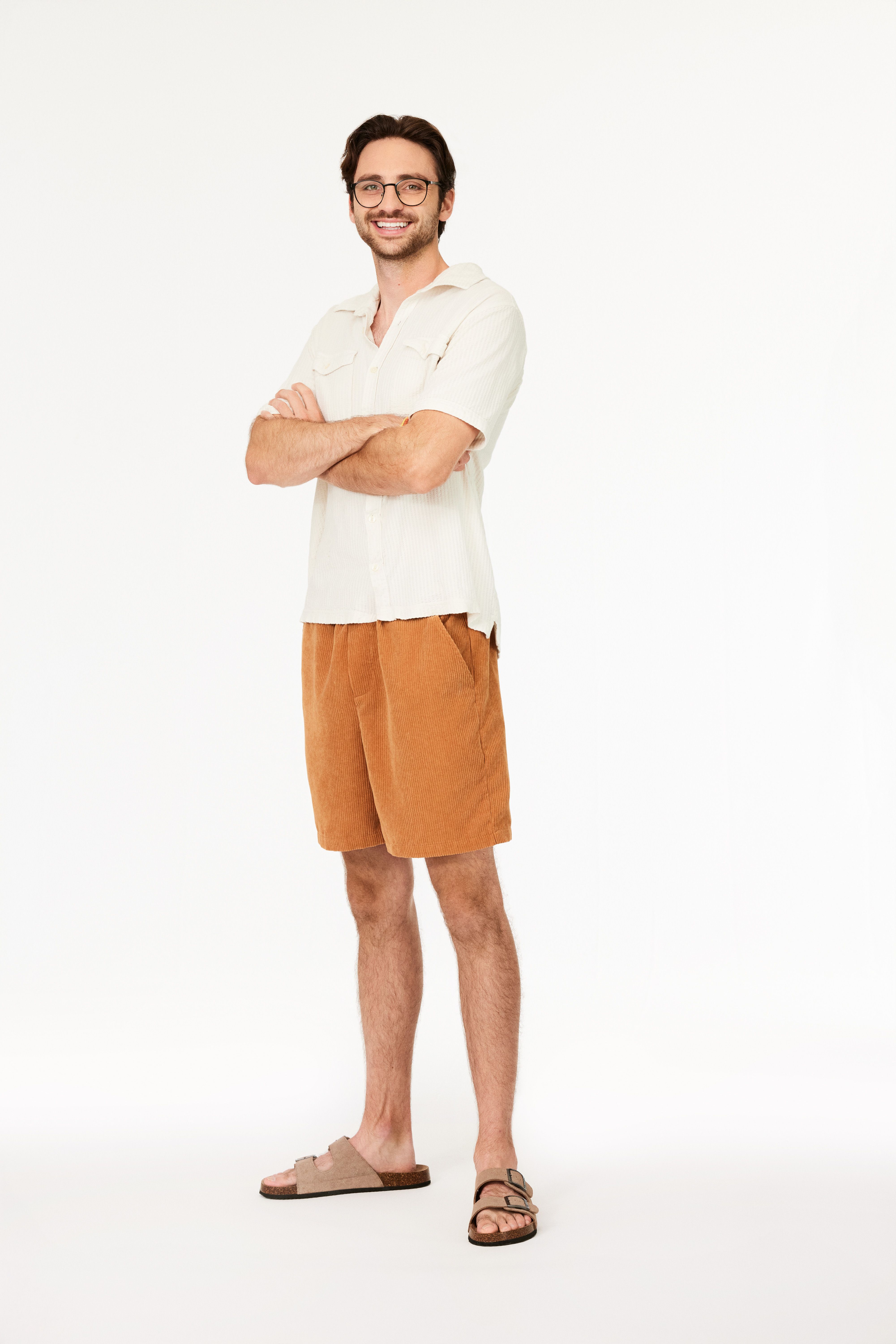 Smiling man with glasses wearing a white short-sleeve shirt, brown corduroy shorts, and beige sandals, standing with arms crossed against a plain white background.