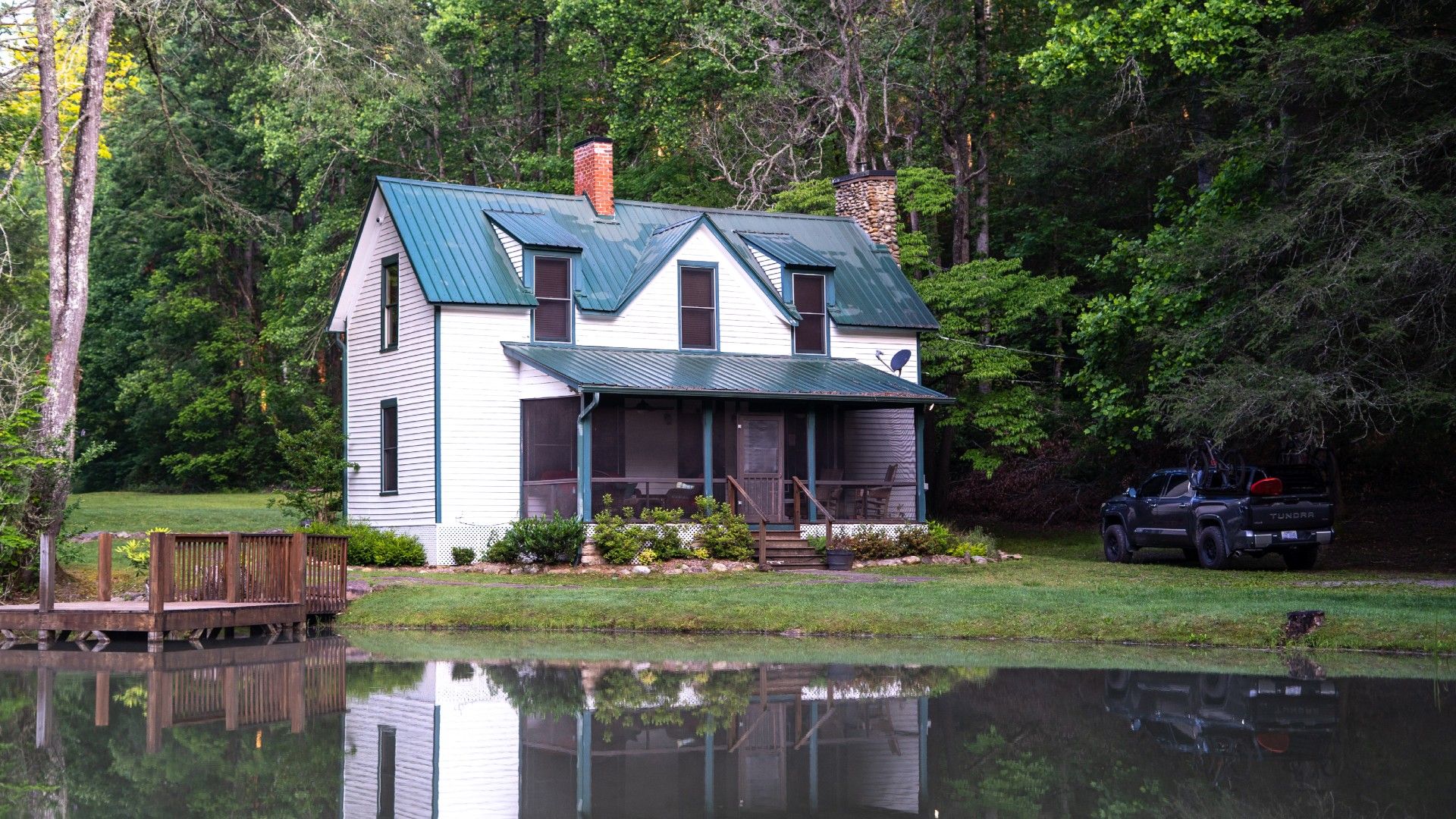 A close-up view of a white lakeside cabin and truck reflected in a calm pond, nestled in dense forest.