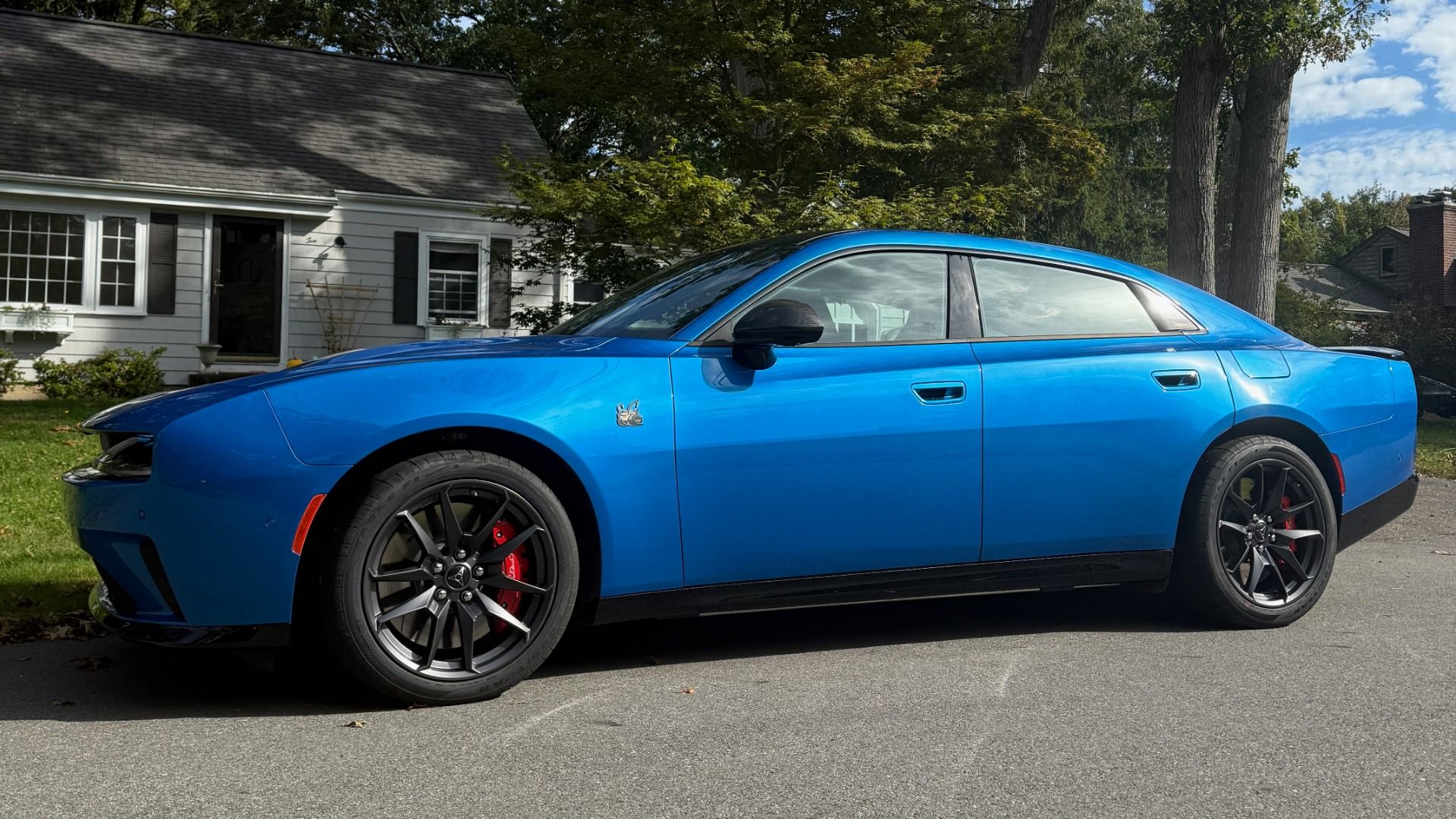 Blue Dodge Charger EV parked on a residential street with black rims and red brake calipers in front of houses and trees.