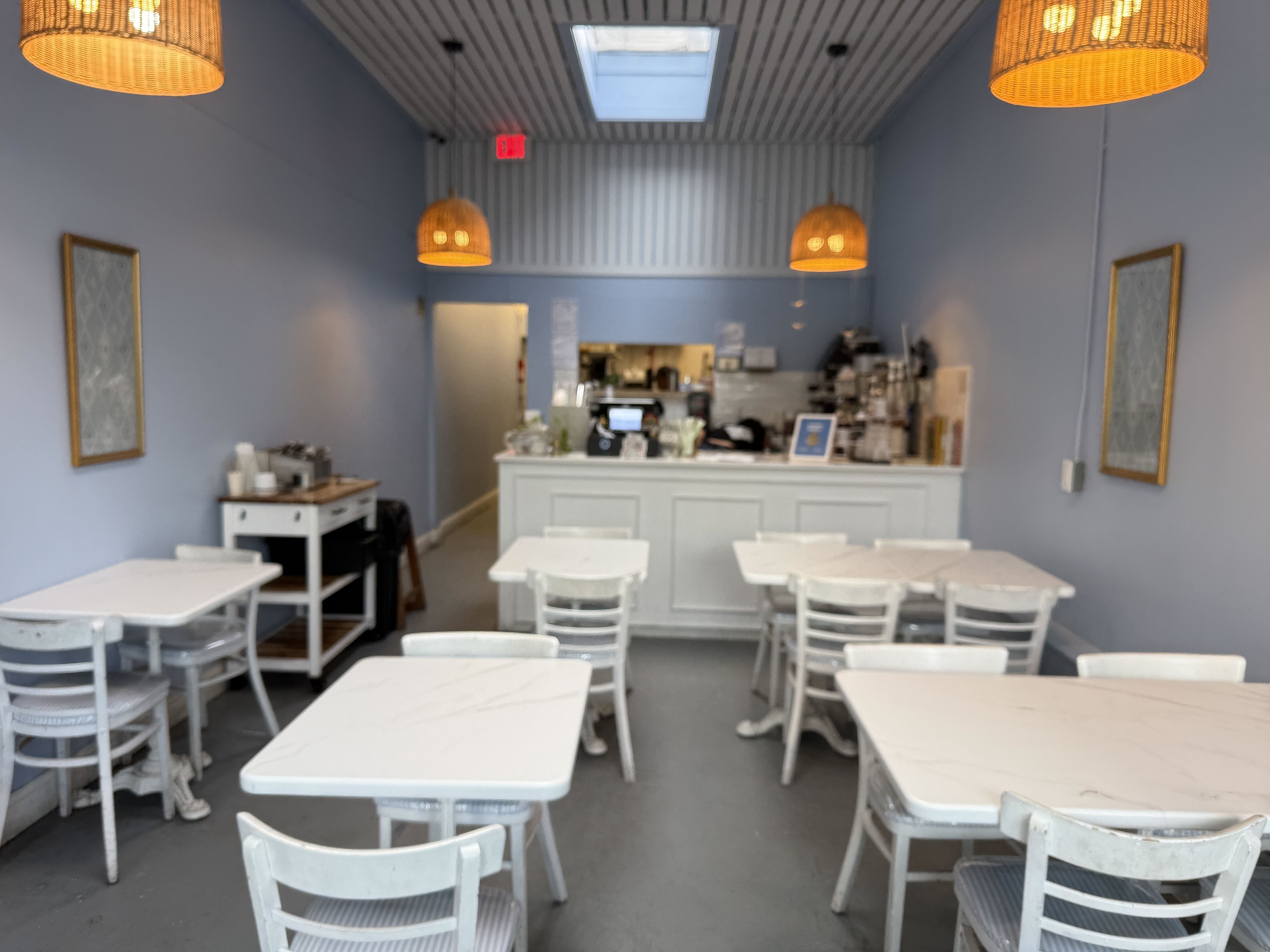 Blue-walled cafe with white marble-topped tables and chairs, wicker pendant lights, and a counter at the back. A red exit sign above a doorway; skylight in the striped ceiling.