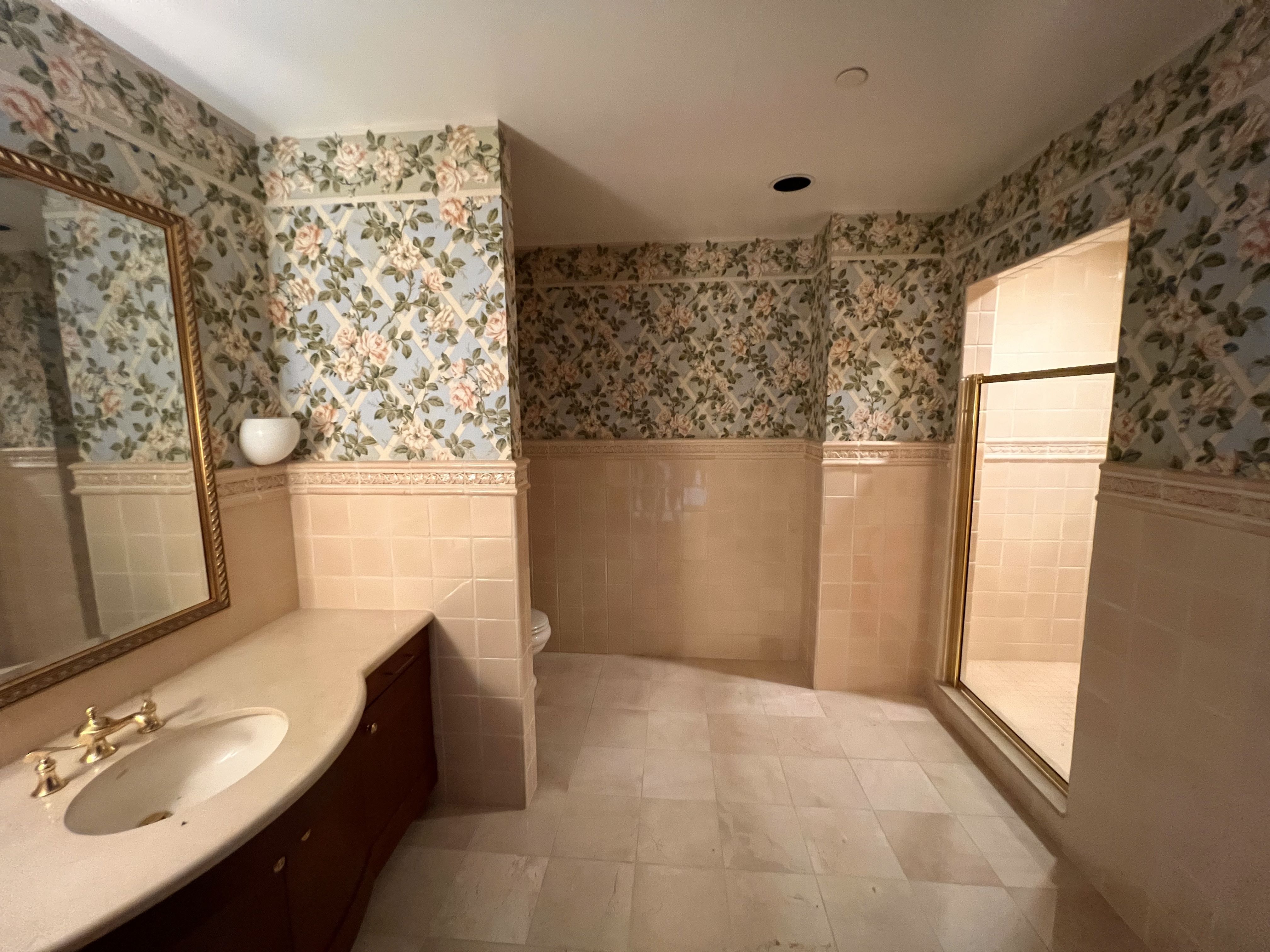 Vintage-style bathroom with blue floral wallpaper and pink roses, beige tile wainscoting. A gold-framed mirror above a cream countertop vanity; toilet behind a wall and a glass shower on the right.