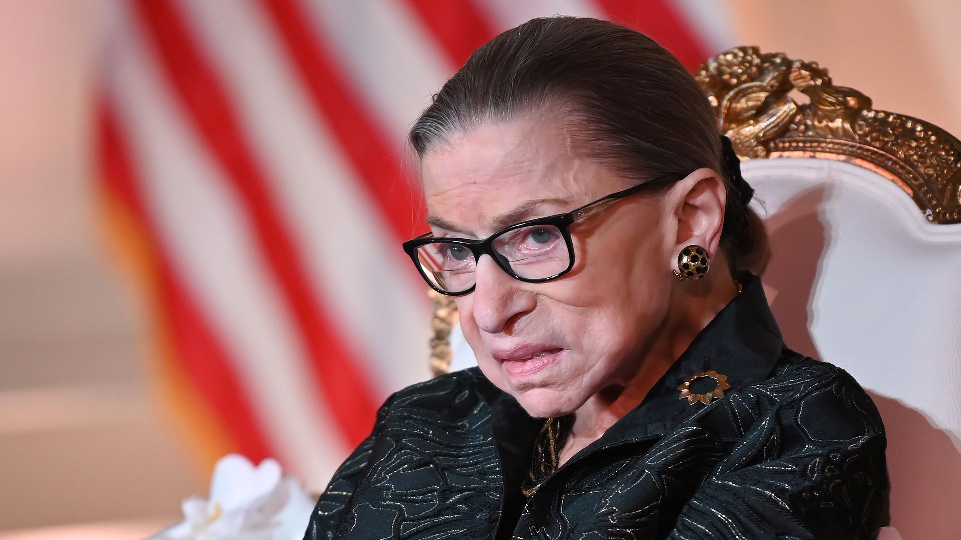 Ruth Bader Ginsburg sits in a white and gold chair with the American flag in the background