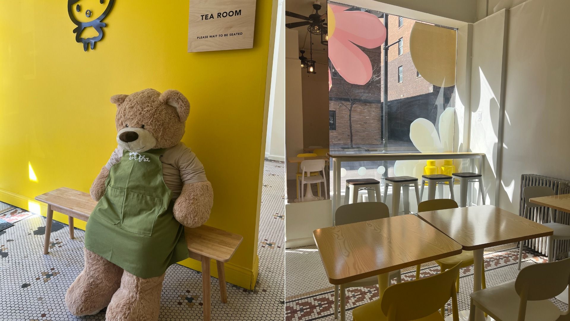 Large teddy bear wearing a green apron sitting on a wooden bench against a yellow wall with a tea room sign. Sunlit cafe interior with wooden tables, chairs, stools, and floral window decals.