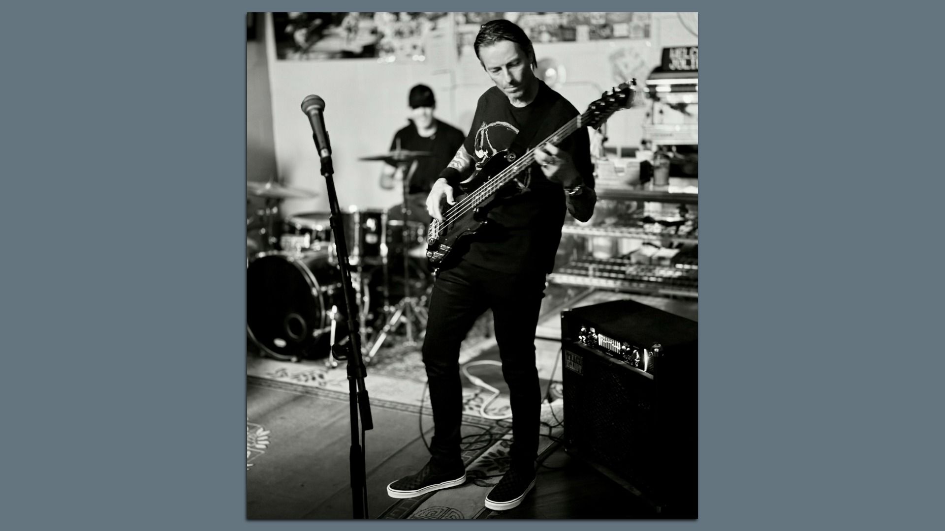 Black and white photo of a musician playing bass guitar in a rehearsal space, with a drum set and microphone stand nearby, and an amplifier on the right side.