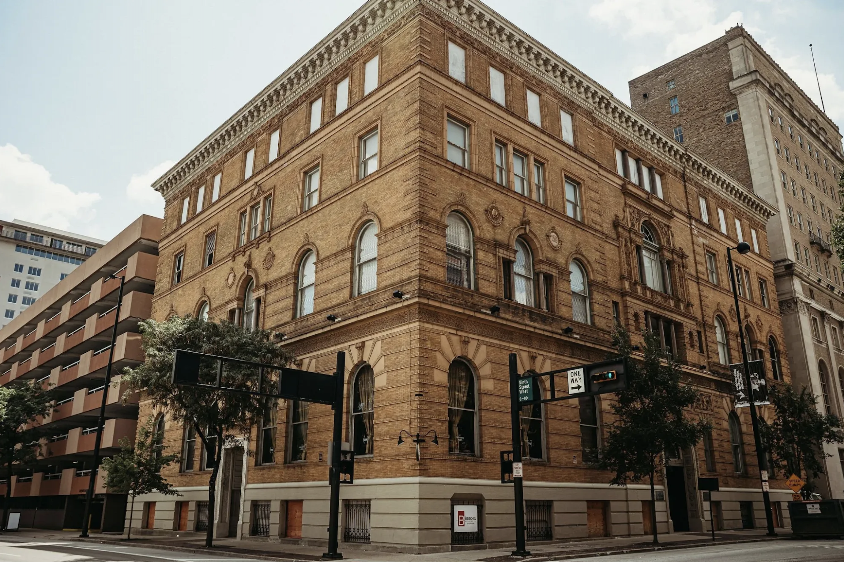 Corner three-story brick building with arched ground-floor windows and decorative stonework along the cornice; a taller brick building towers behind at a busy city intersection under a blue sky.