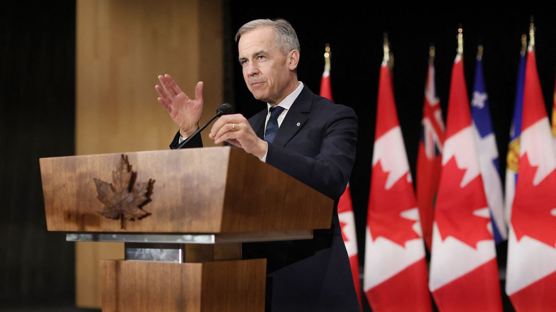 Mark Carney speaks behind a podium decorated with a Maple leaf in front of several Canadian flags.