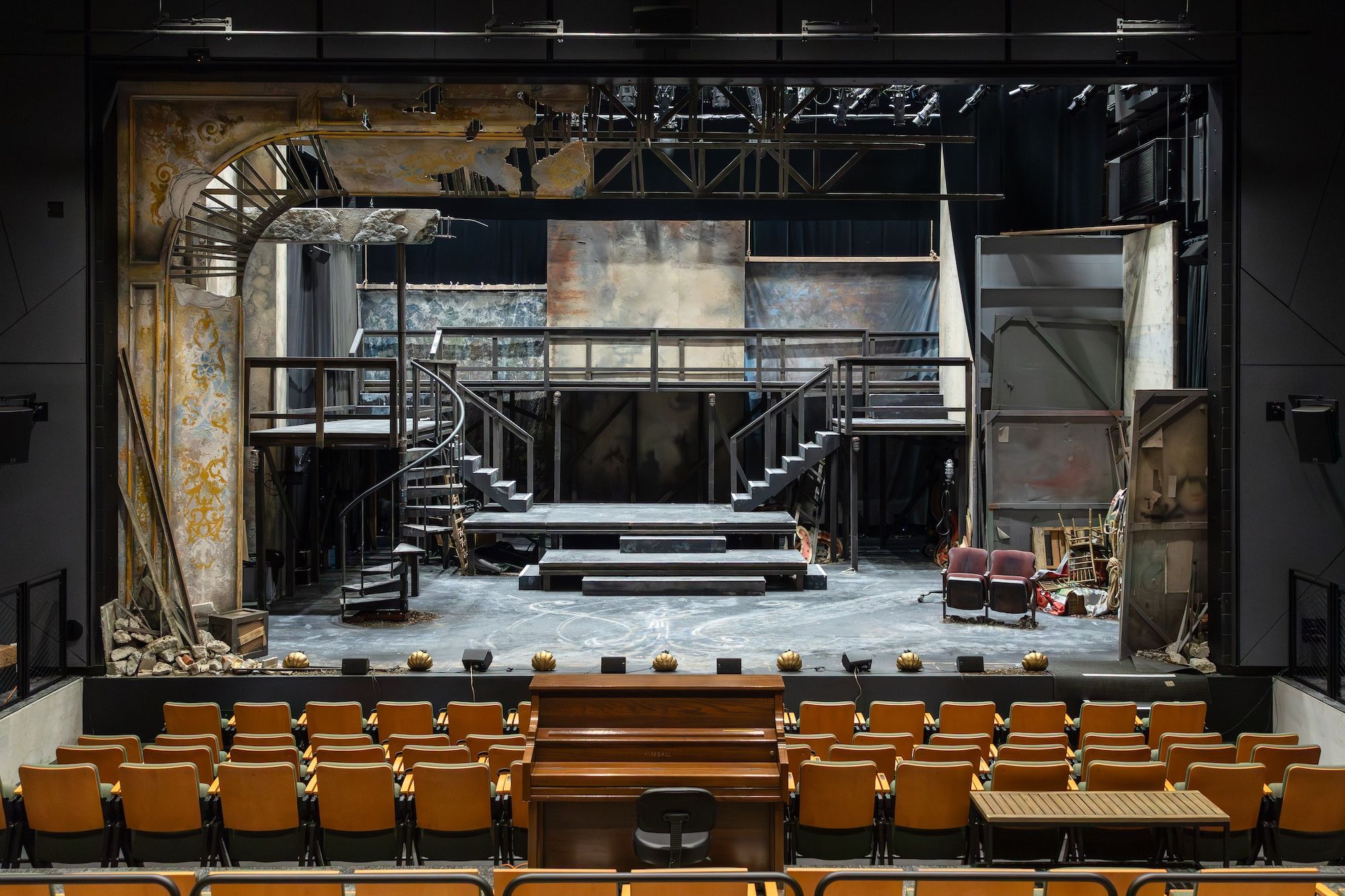 Empty theater stage with a rustic, weathered set featuring metal stairs, platforms, and faded backdrops. Yellow seating and a piano are visible in the foreground under dim lighting.