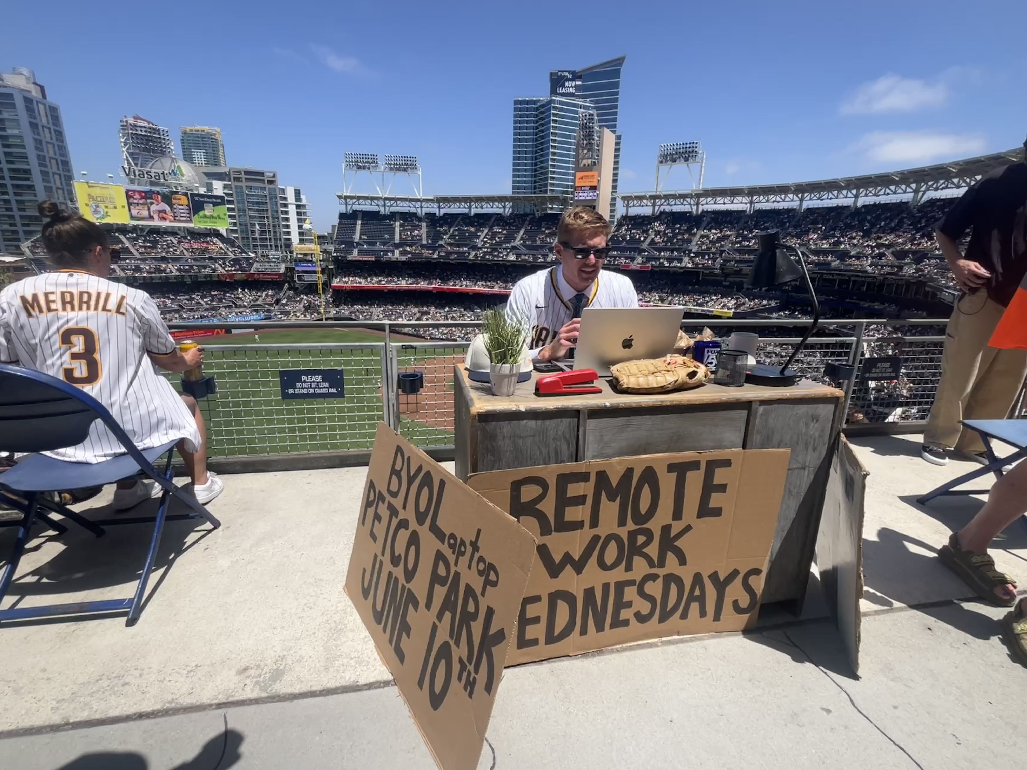 Sunny baseball stadium with empty seats. A man in a white pinstripe jersey sits at a desk with a laptop and glove; cardboard signs read "REMOTE WORK WEDNESDAYS" in the foreground as another person stands nearby.