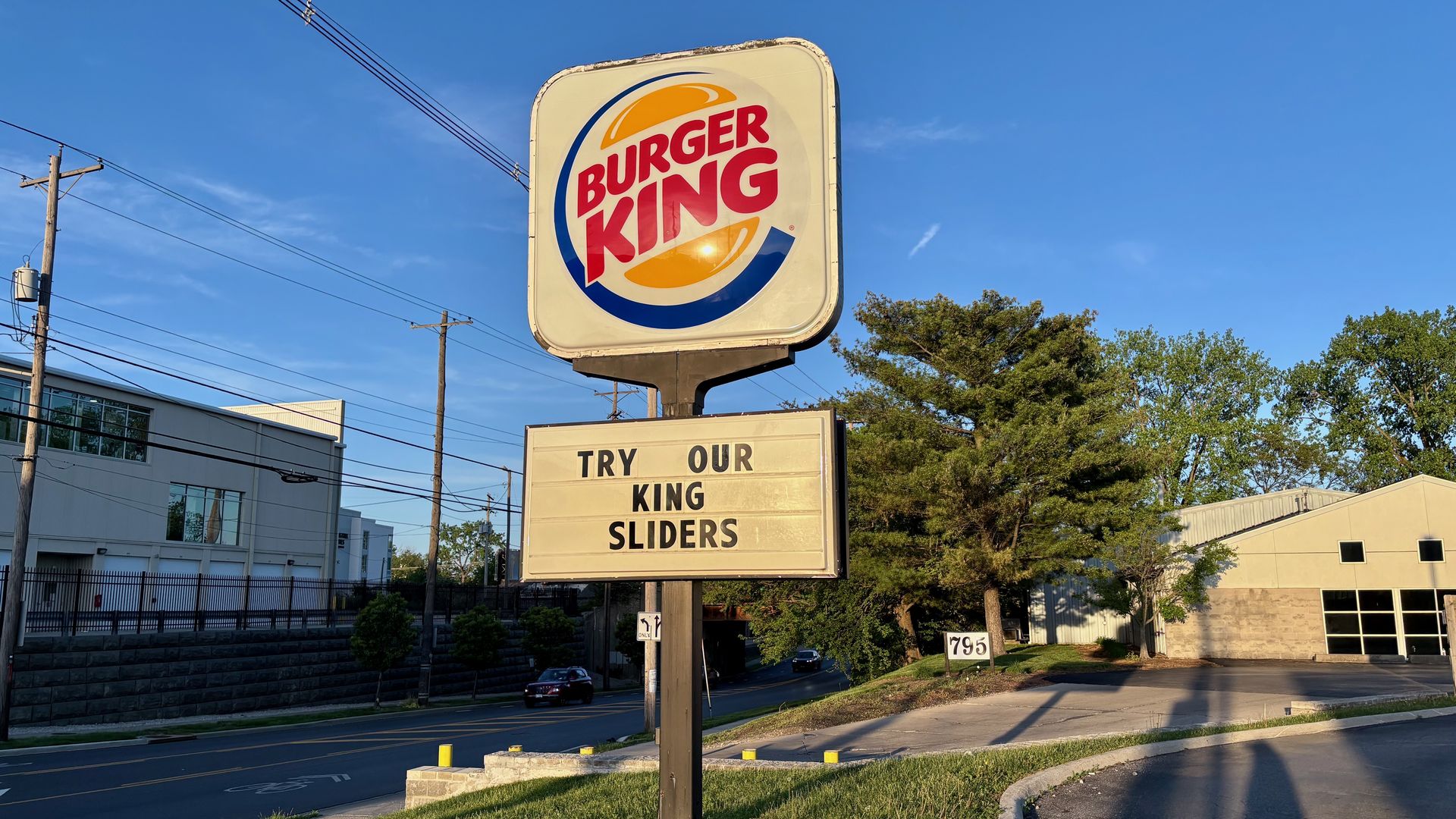 Burger King sign with red logo inside a blue and yellow circle, set against a clear blue sky. A roadside marquee reads TRY OUR KING SLIDERS, with trees and buildings in the background.