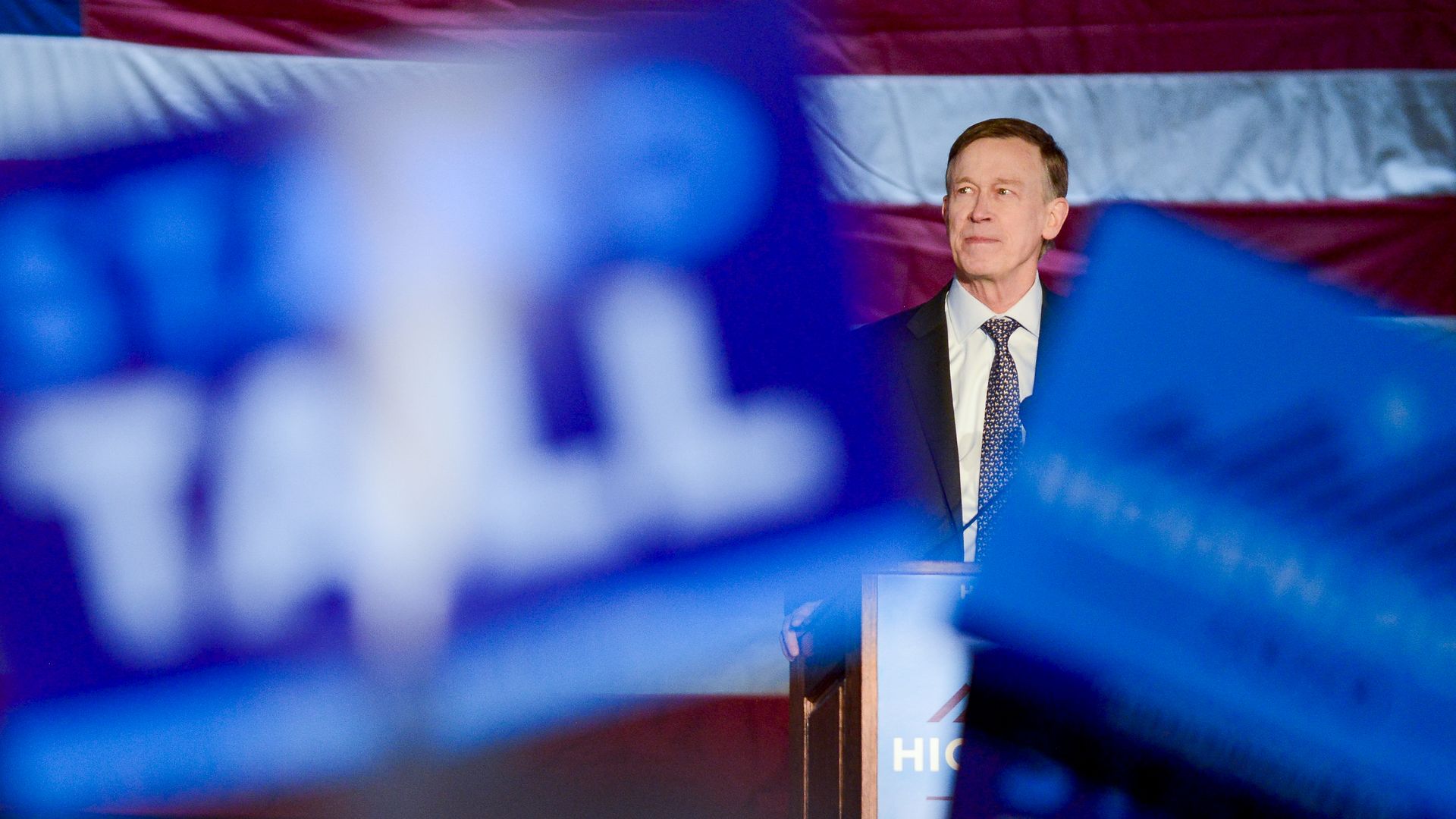 Hickenlooper speaks from behind a podium at a campaign rally, framed by blue signs. 