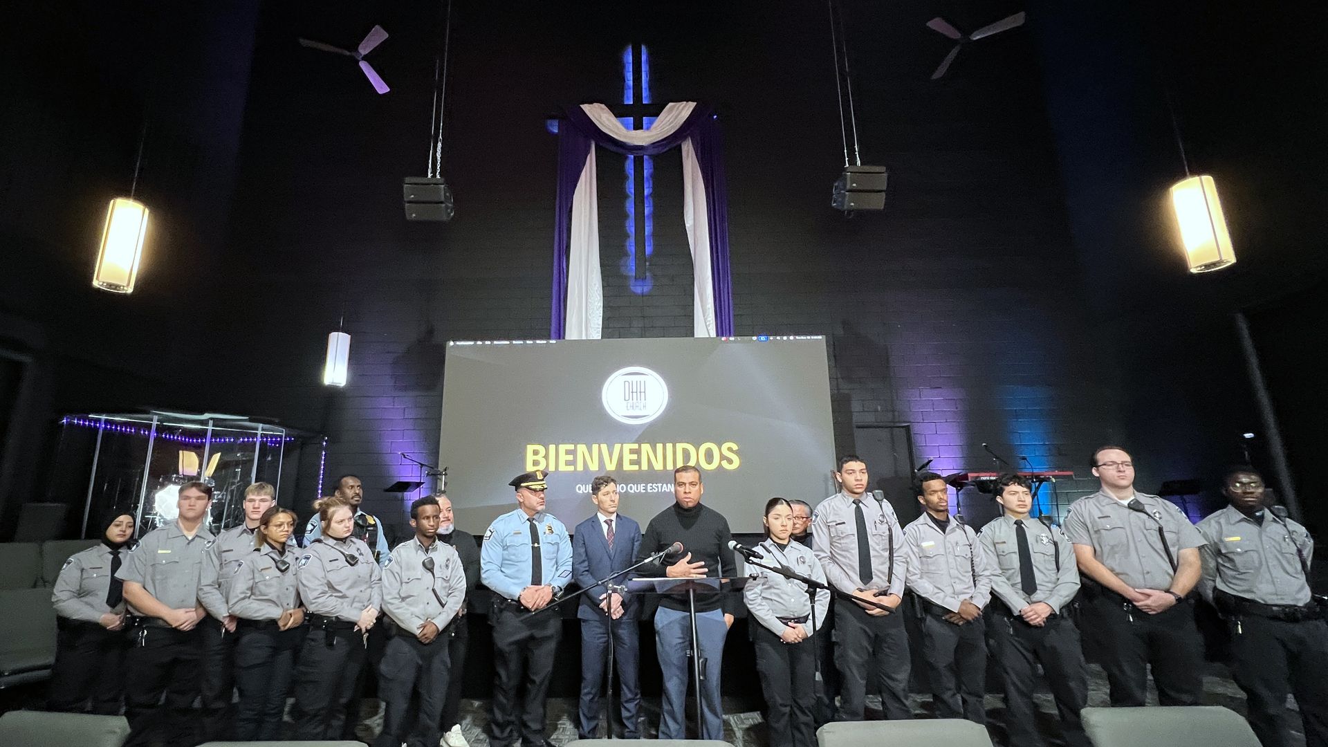 Group of uniformed security or police officers and two men in suits standing in a row inside a dark room with a cross and purple-white drapes above a screen reading "BIENVENIDOS".