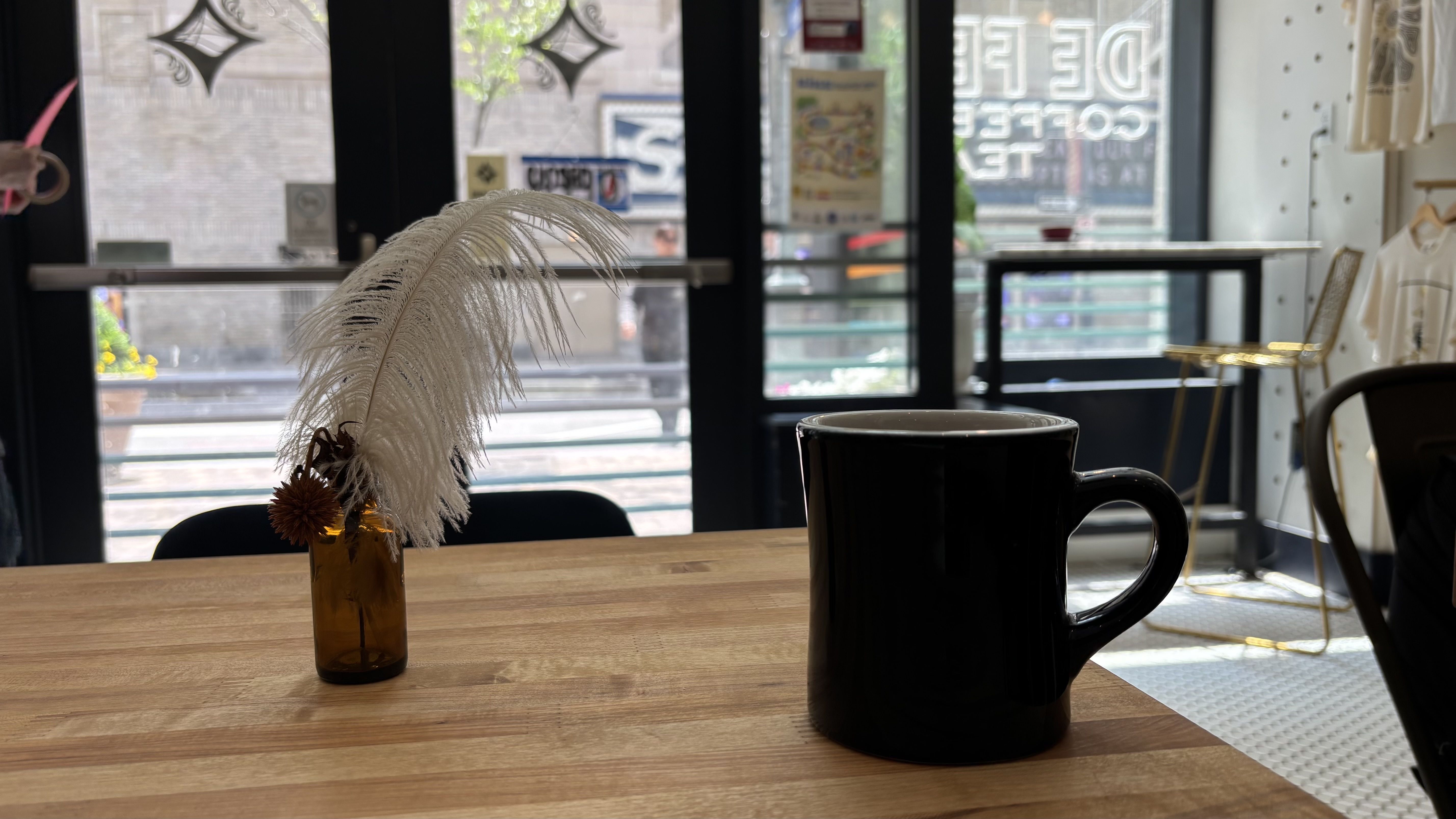 Inside a cafe, an amber vase with white dried feathers sits on a wooden table, a large black mug in the foreground. Glass doors behind reveal a street scene outside.