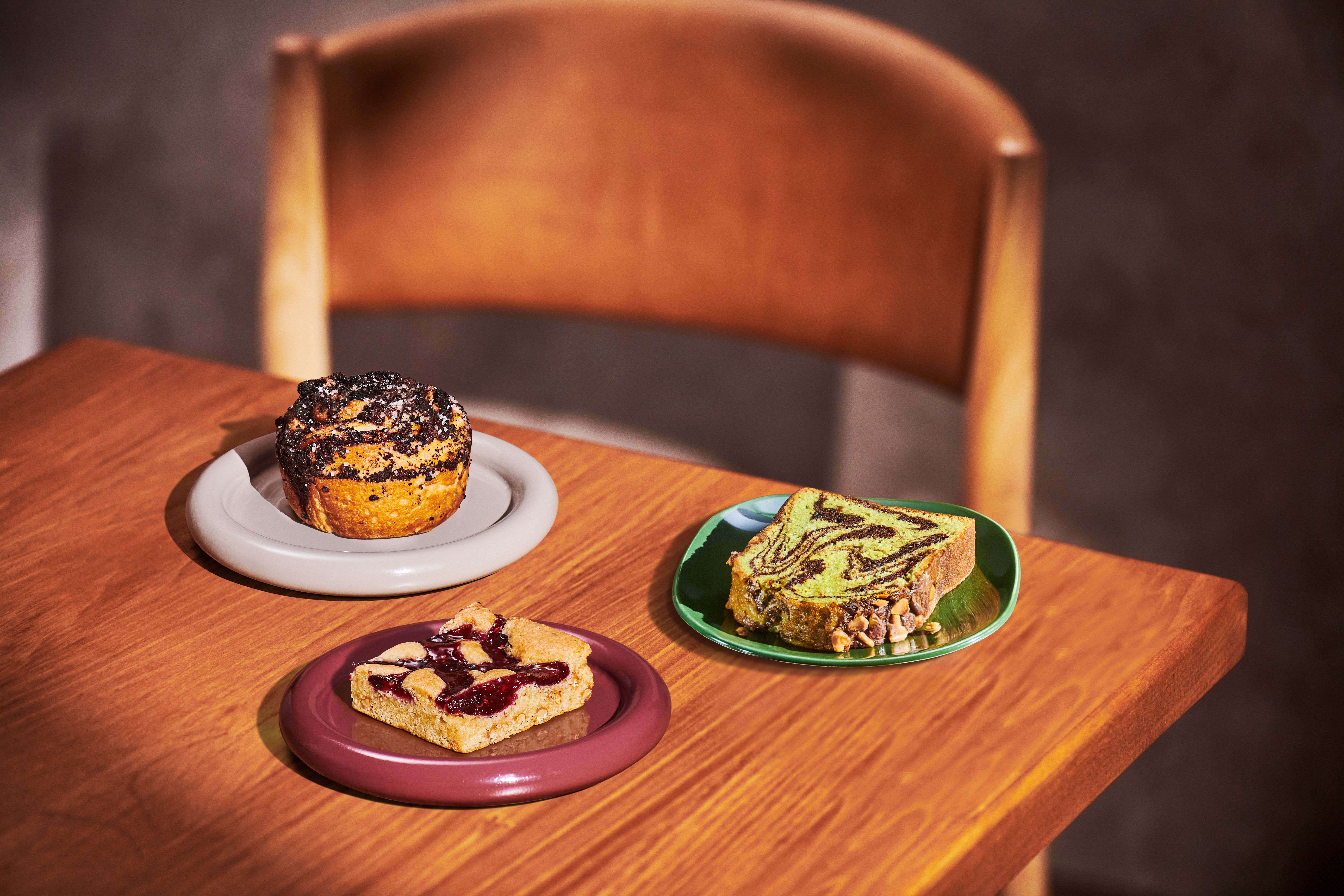 Three baked treats on colorful plates on a wooden table: a crumbly muffin on a gray plate, a fruit-topped bar on a purple plate, and a green-pistachio swirled cake slice on a green plate, with a wooden chair in the background.