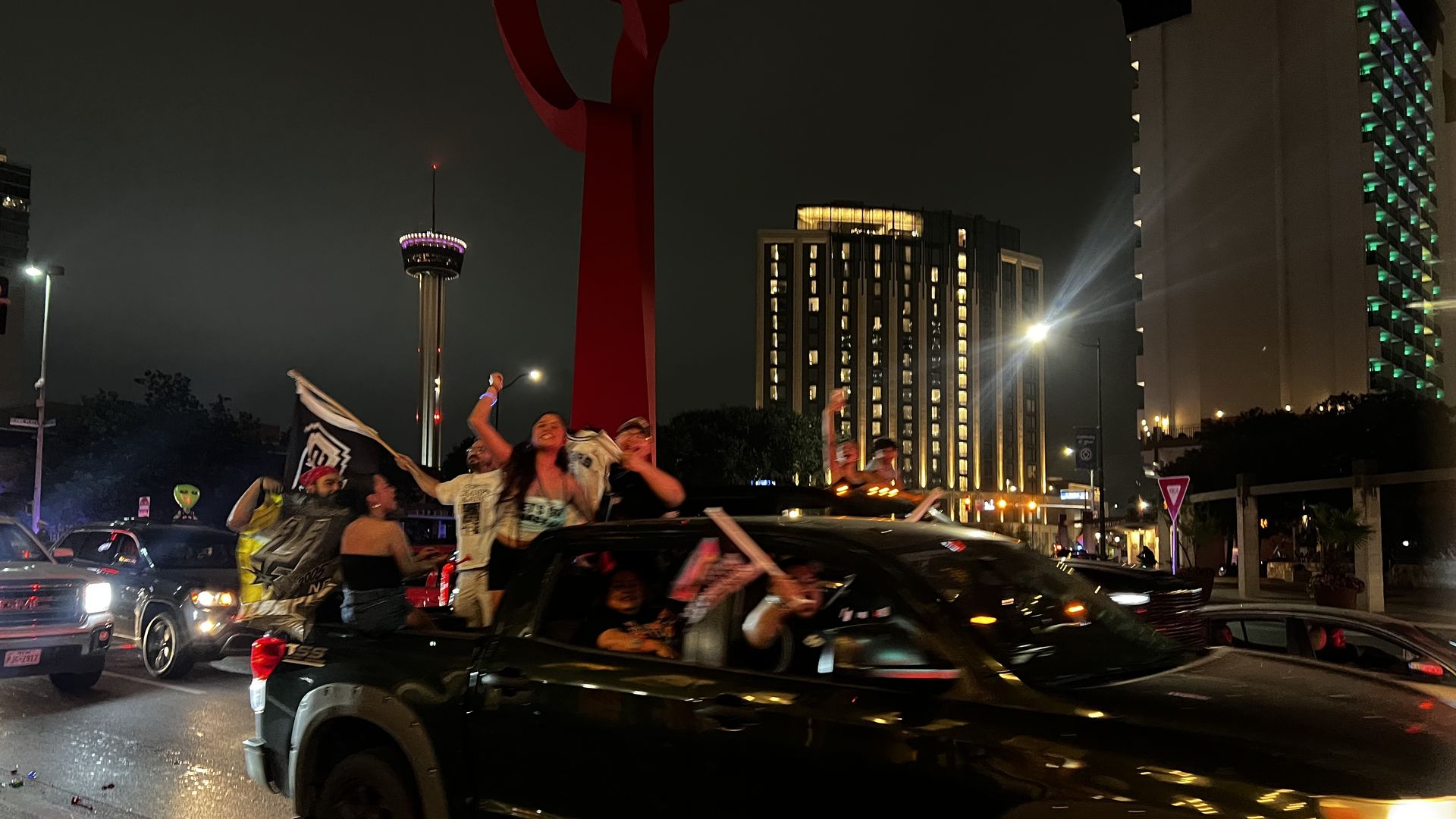 Nighttime city street with a parade in a convertible, confetti flying. A tall crimson sculpture dominates the foreground while illuminated buildings and a Hilton hotel tower rise in the background.