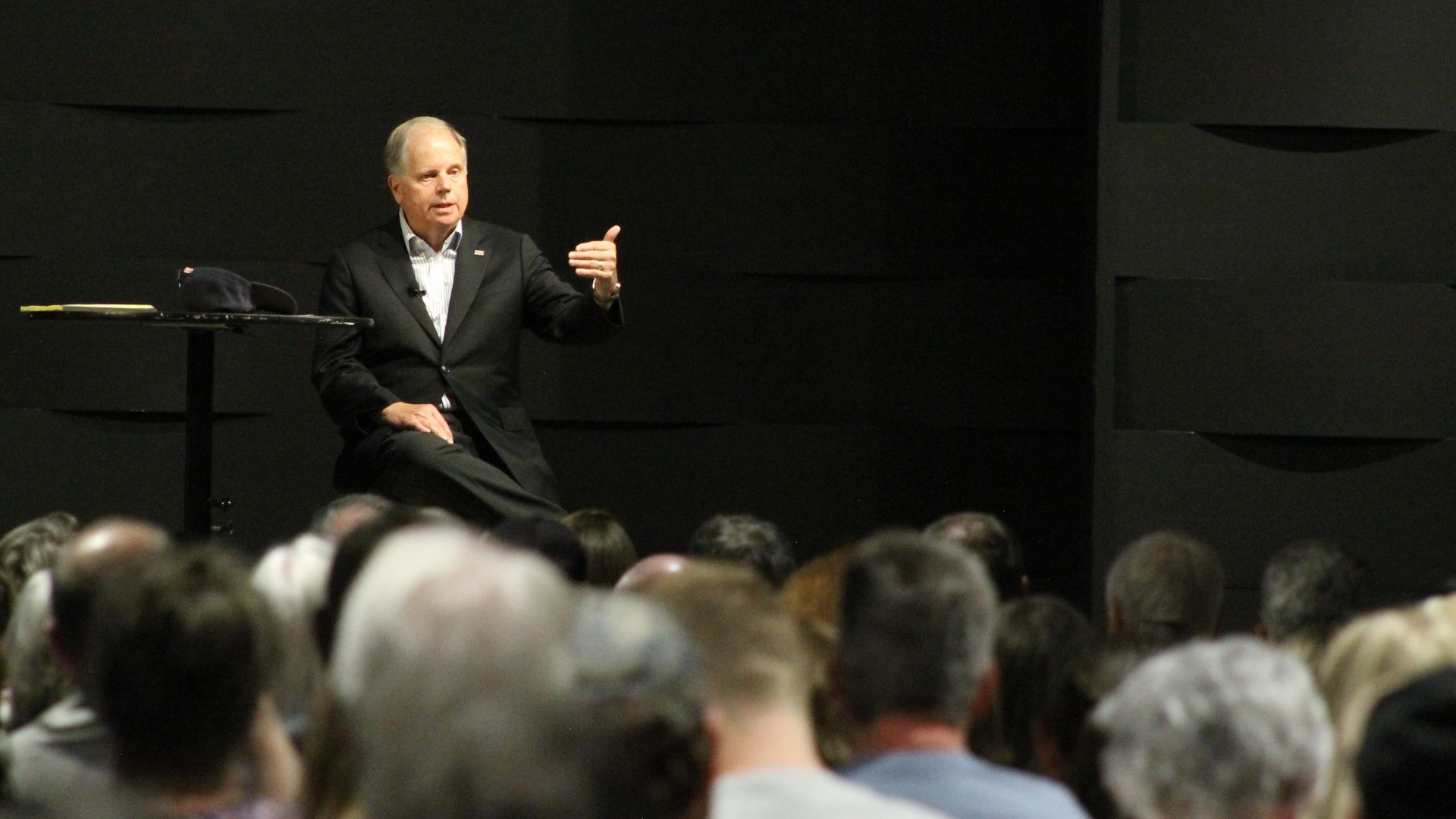 A middle-aged man in a dark suit sits on a stage, gesturing with his right hand as he speaks to an audience; a table with a hat and notebook sits nearby against a black paneled backdrop.