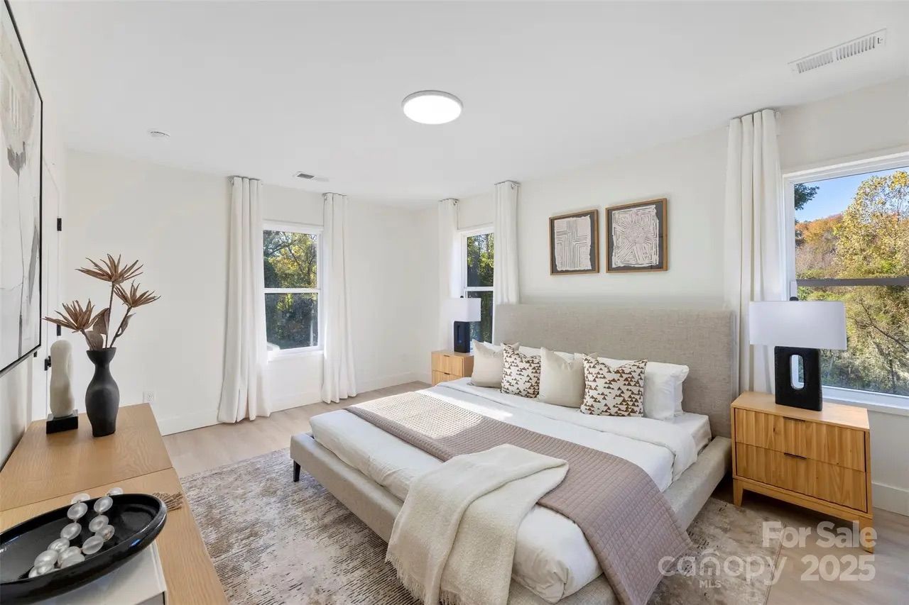 Bright bedroom with white walls, beige upholstered bed, patterned pillows, light wood nightstands, black modern lamps, large windows with white curtains, and decorative vases on dresser.