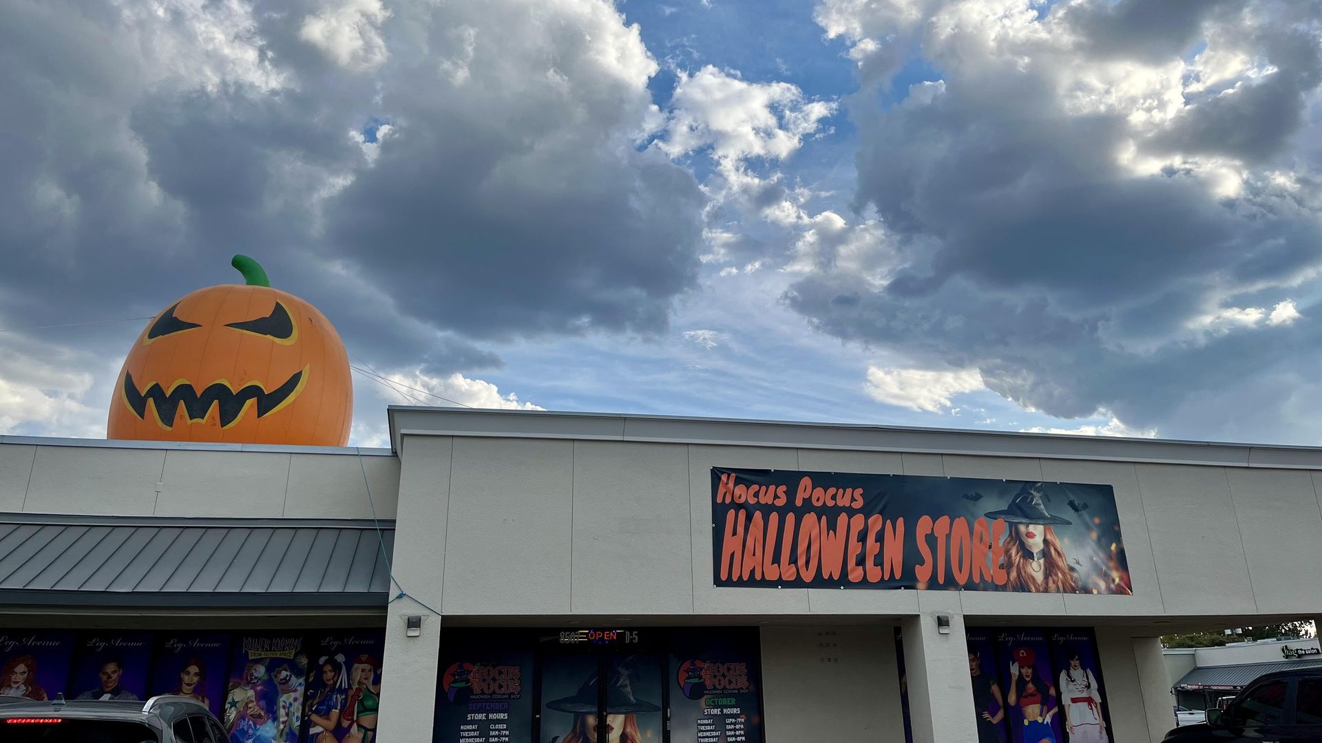 A building with a large inflatable scary pumpkin on the roof and a sign reading "Hocus Pocus Halloween Store" against a dramatic cloudy blue sky.