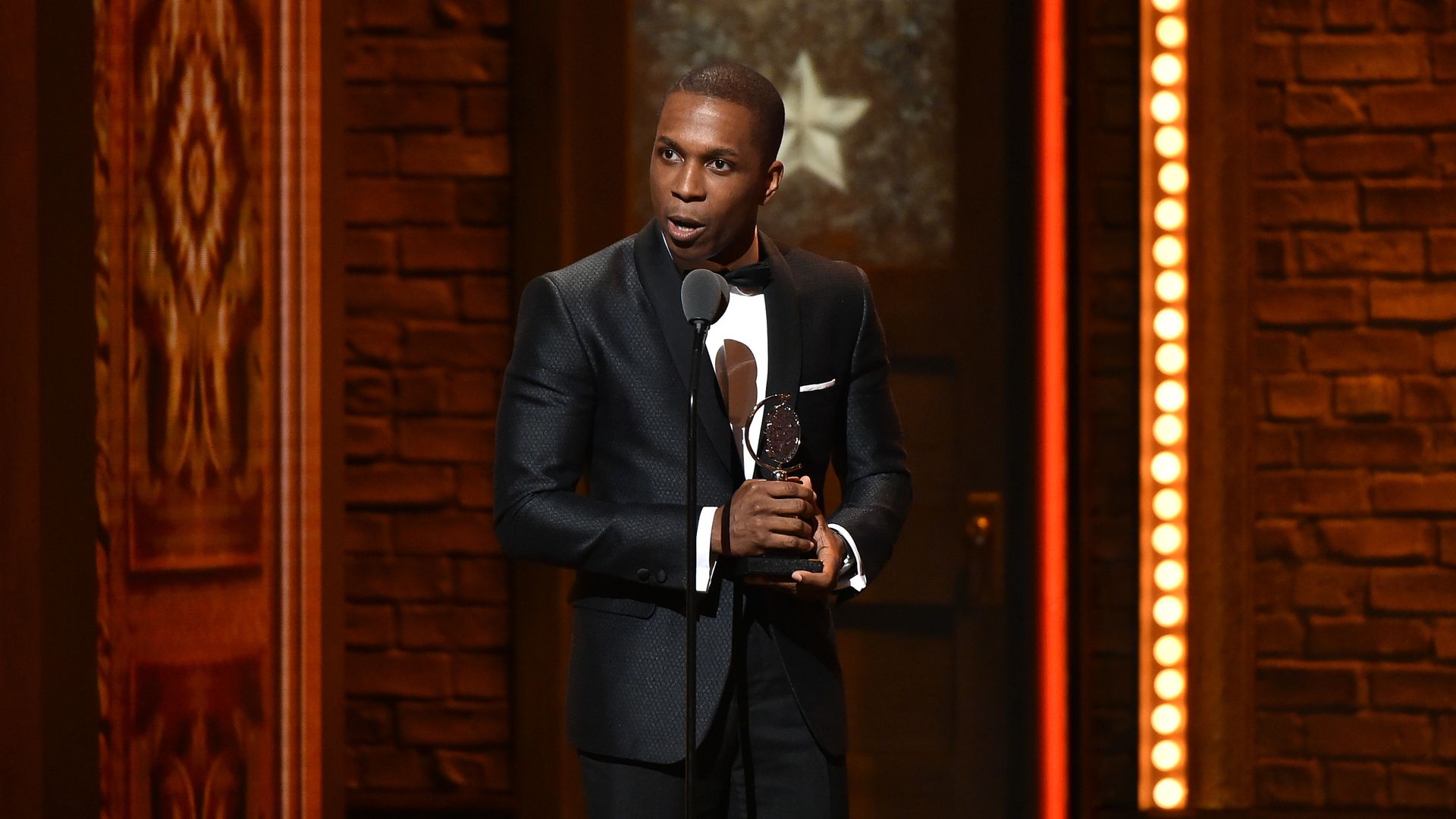 A person holds a golden statuette while taking into a microphone and wearing a tuxedo