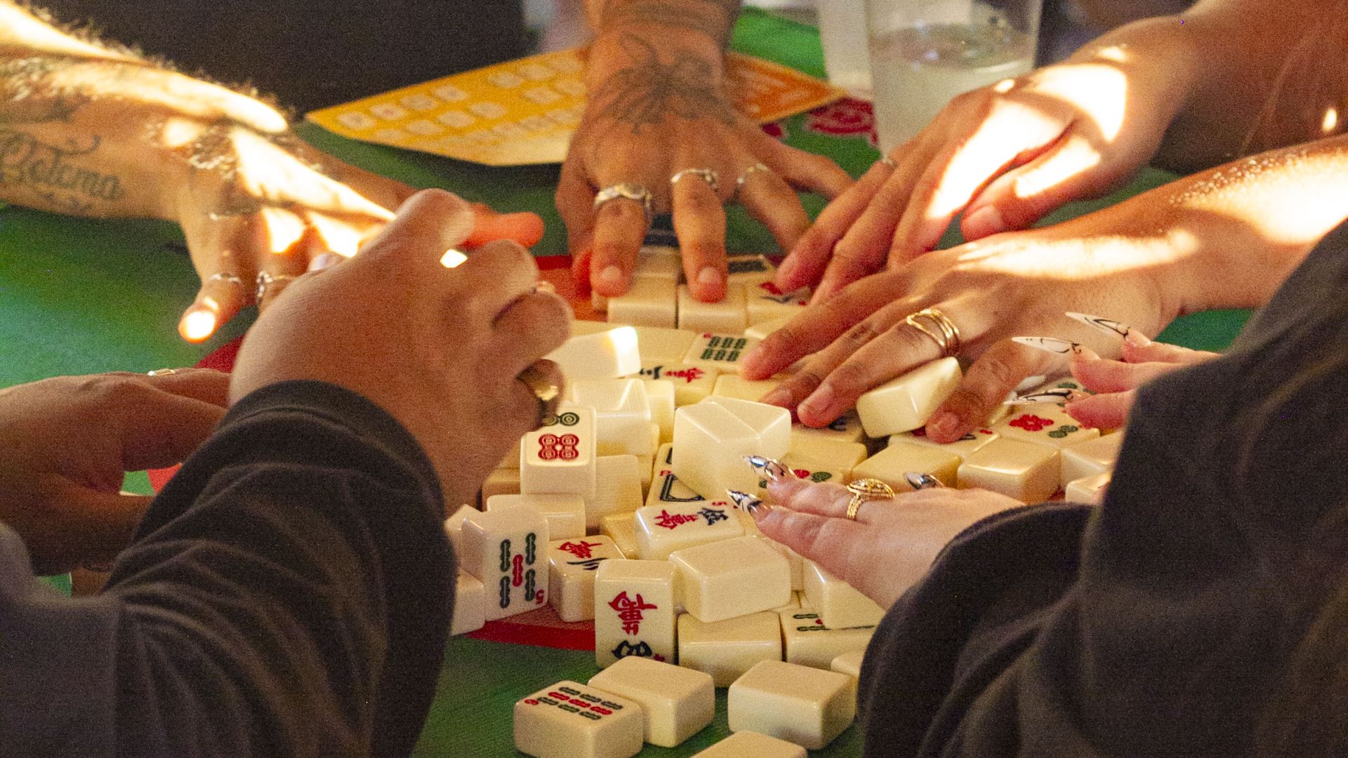 Several people with tattooed and ring-adorned hands playing mahjong on a green table cloth with red patterns, surrounded by white tiles and two clear plastic cups with water.