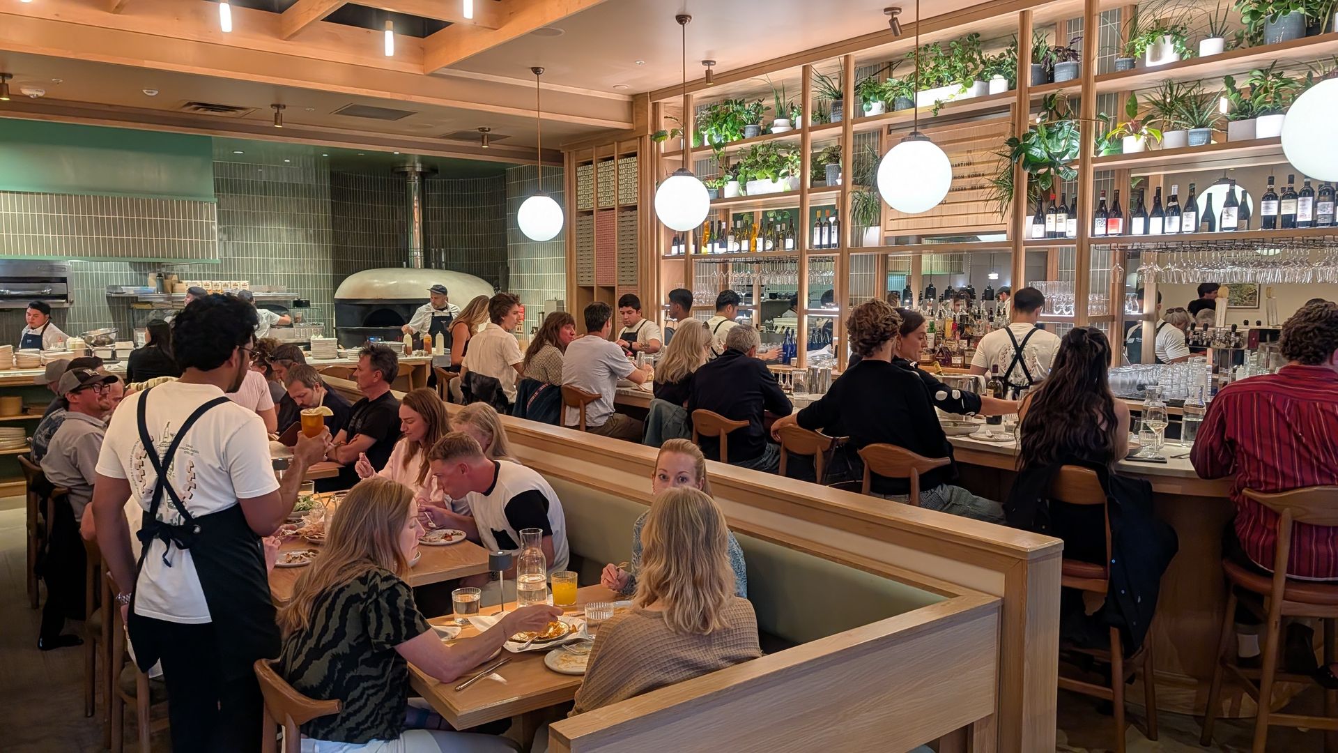 Busy restaurant interior with a long wooden bar, shelves of bottles and hanging plants. Diners sit at tables and stools while chefs work near a large pizza oven in the open kitchen.