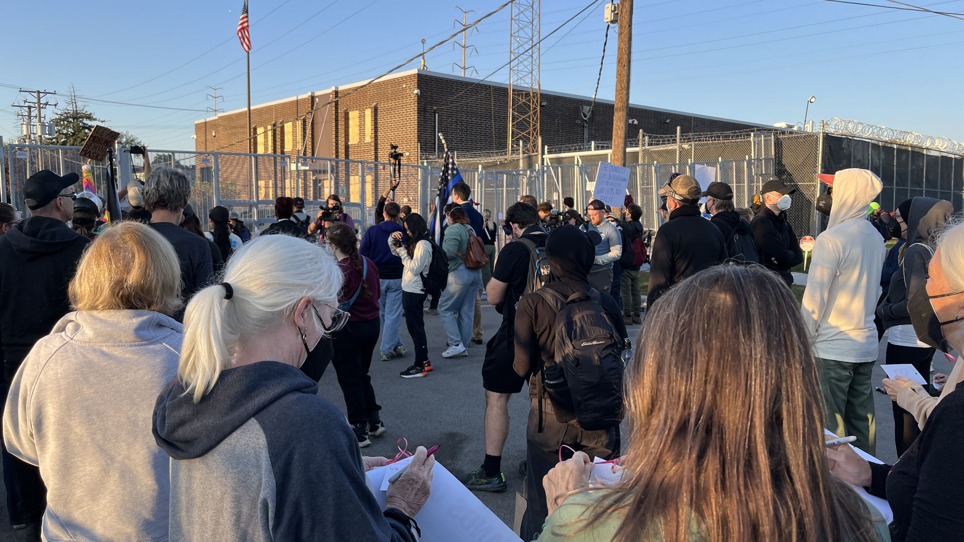 Crowd of masked protesters gathered outside a fenced, secured building with police presence, holding signs and an American flag on a pole in clear daylight.