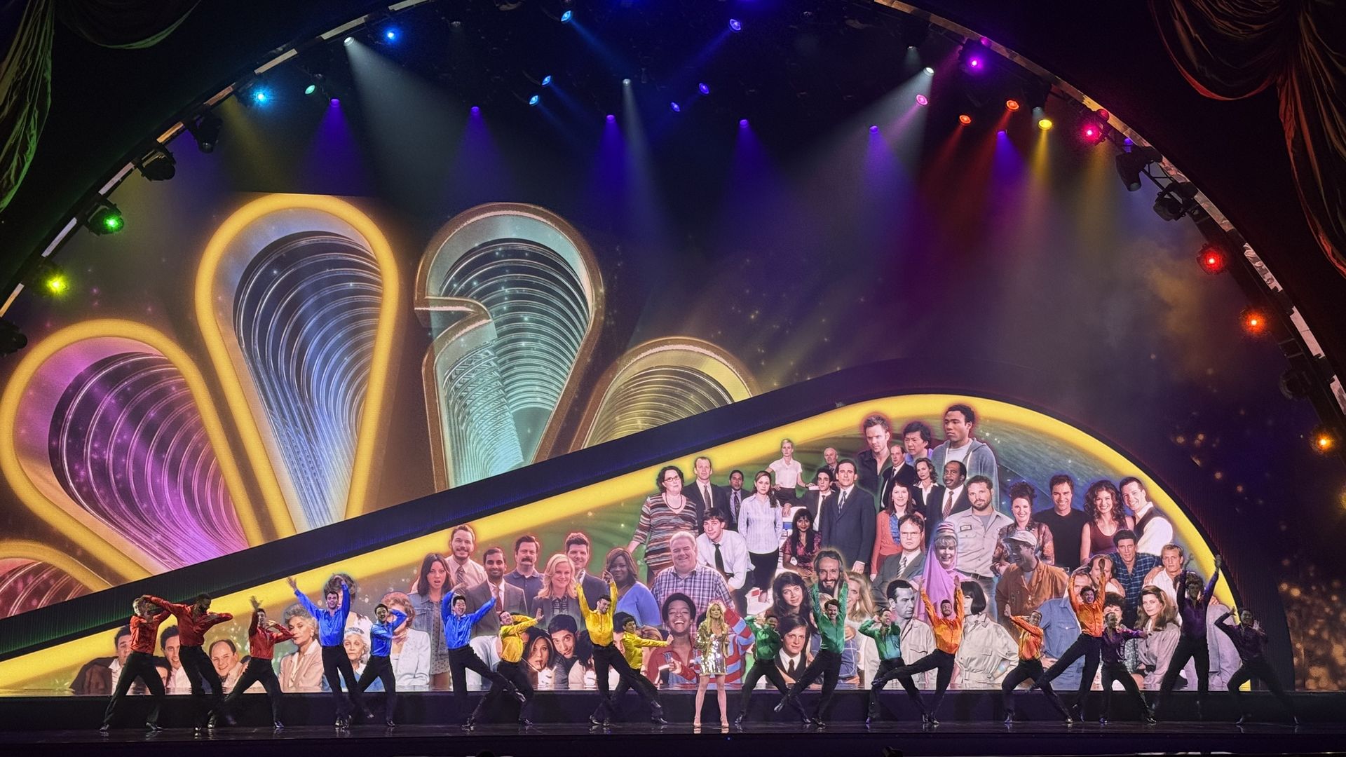 A theater stage with vibrant rainbow arches and concentric patterns, illuminated by colorful overhead lights. A large cast photo banner curves along the back, while dancers perform at the front.
