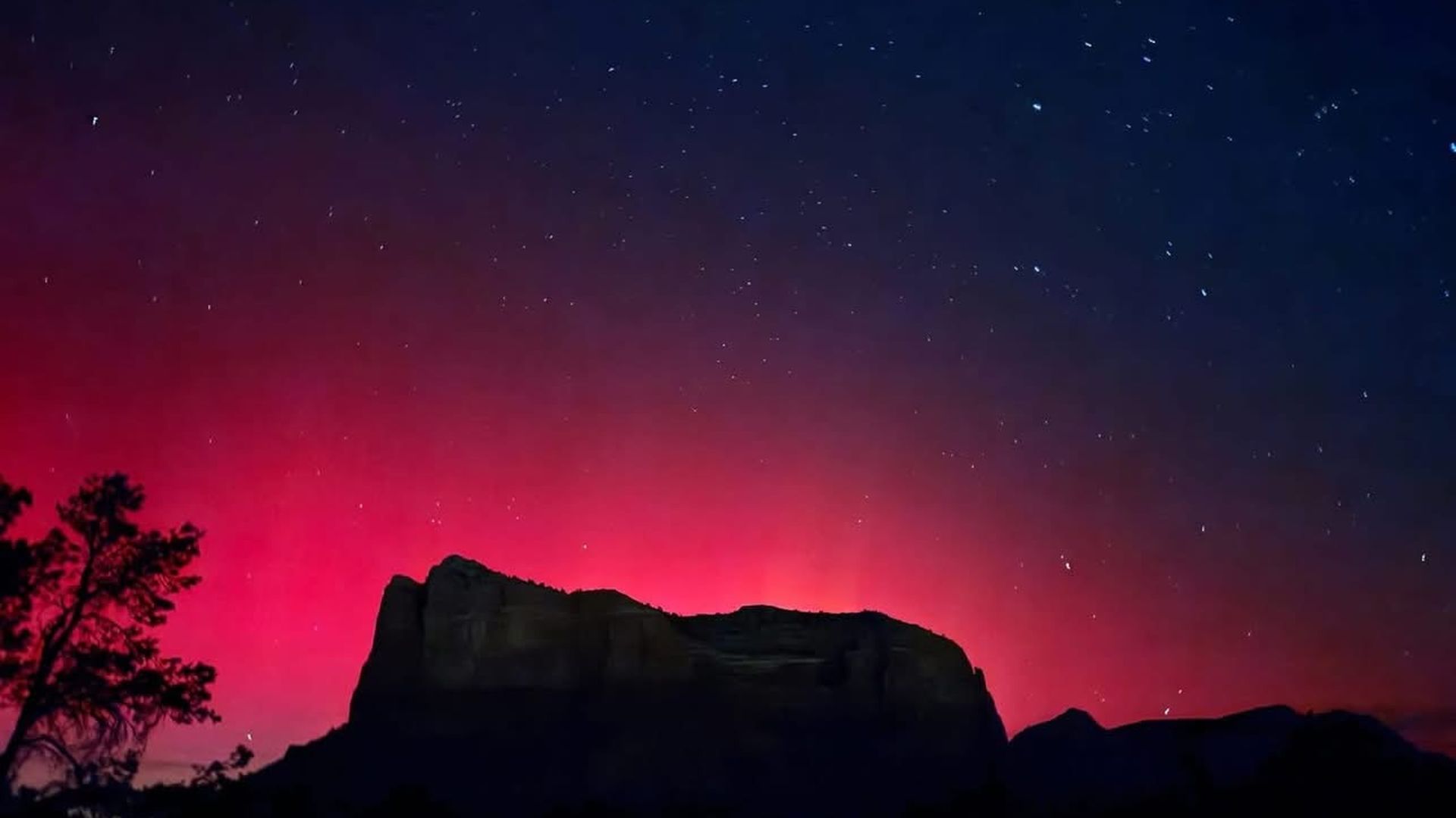 Silhouetted mountain and tree under a starry night sky with a vibrant red aurora or glow behind the mountain.