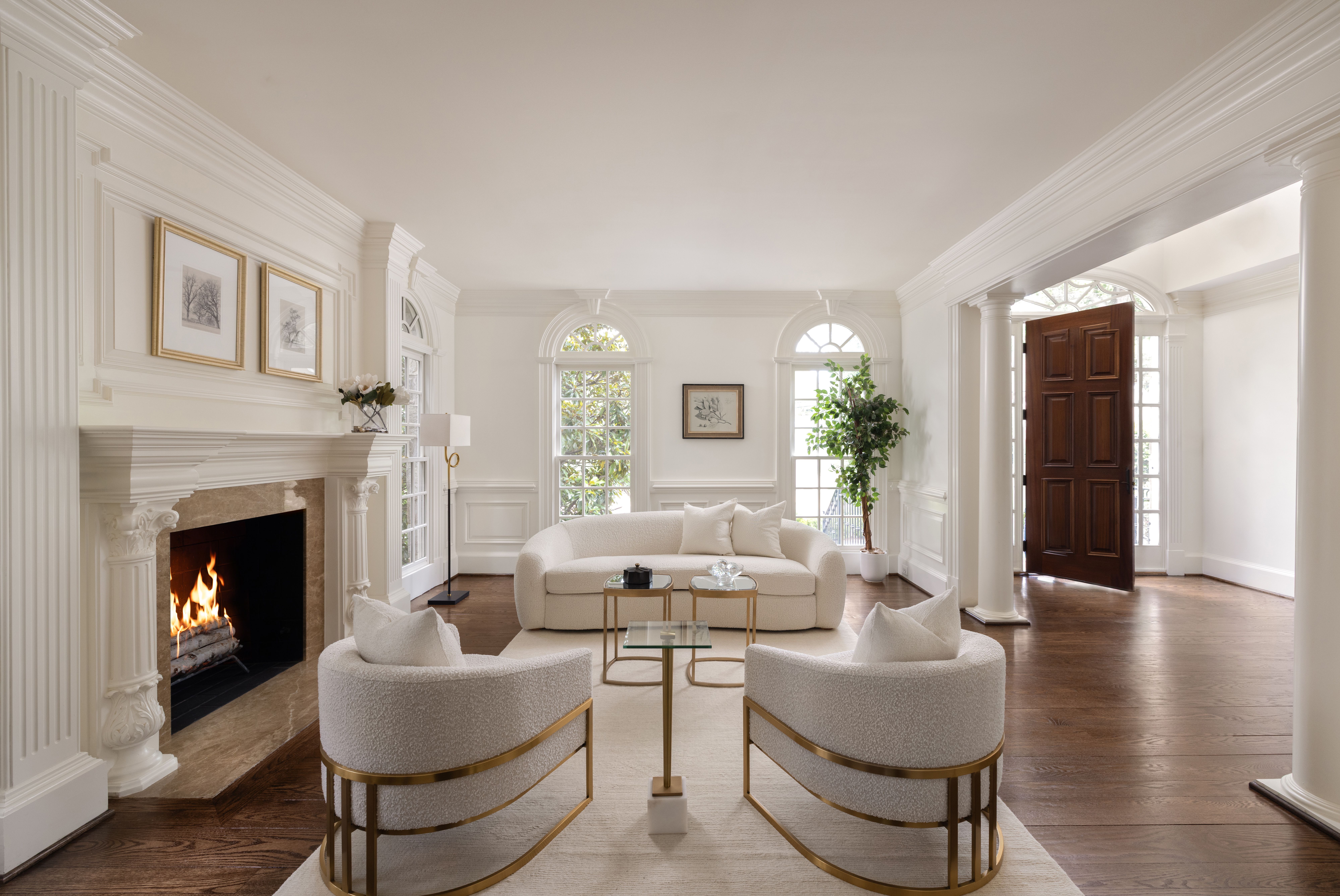Bright living room with white walls, ornate molding, and a lit fireplace. Cream curved sofa set with gold metal frames, glass coffee table, arched windows, a potted plant, and a dark wood front door.