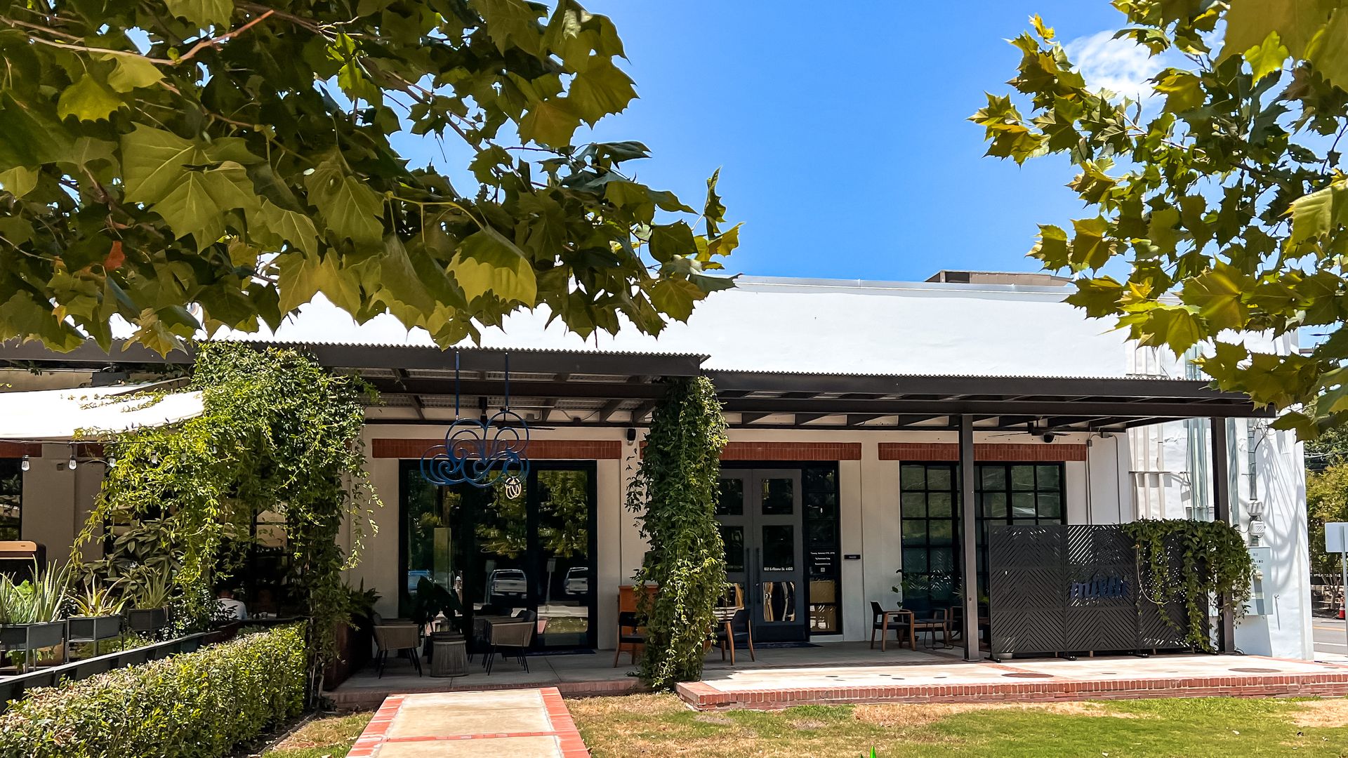 A white building with patio seating and leafy trees.