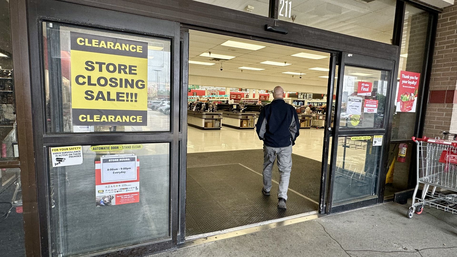 Man walking into a store with glass automatic doors, large yellow clearance signs saying "STORE CLOSING SALE!!!", and visible checkout counters inside.