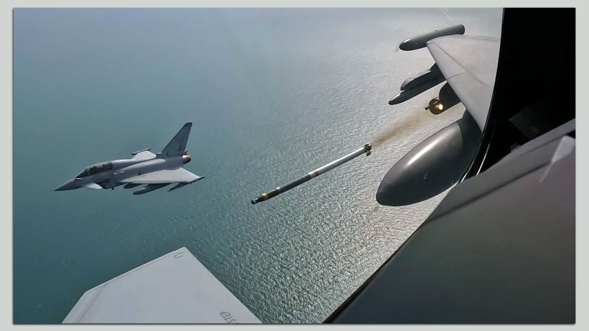 Two gray fighter jets fly over the ocean, viewed from a cockpit window. One is on the left, at distance, while a second is closer on the right side.