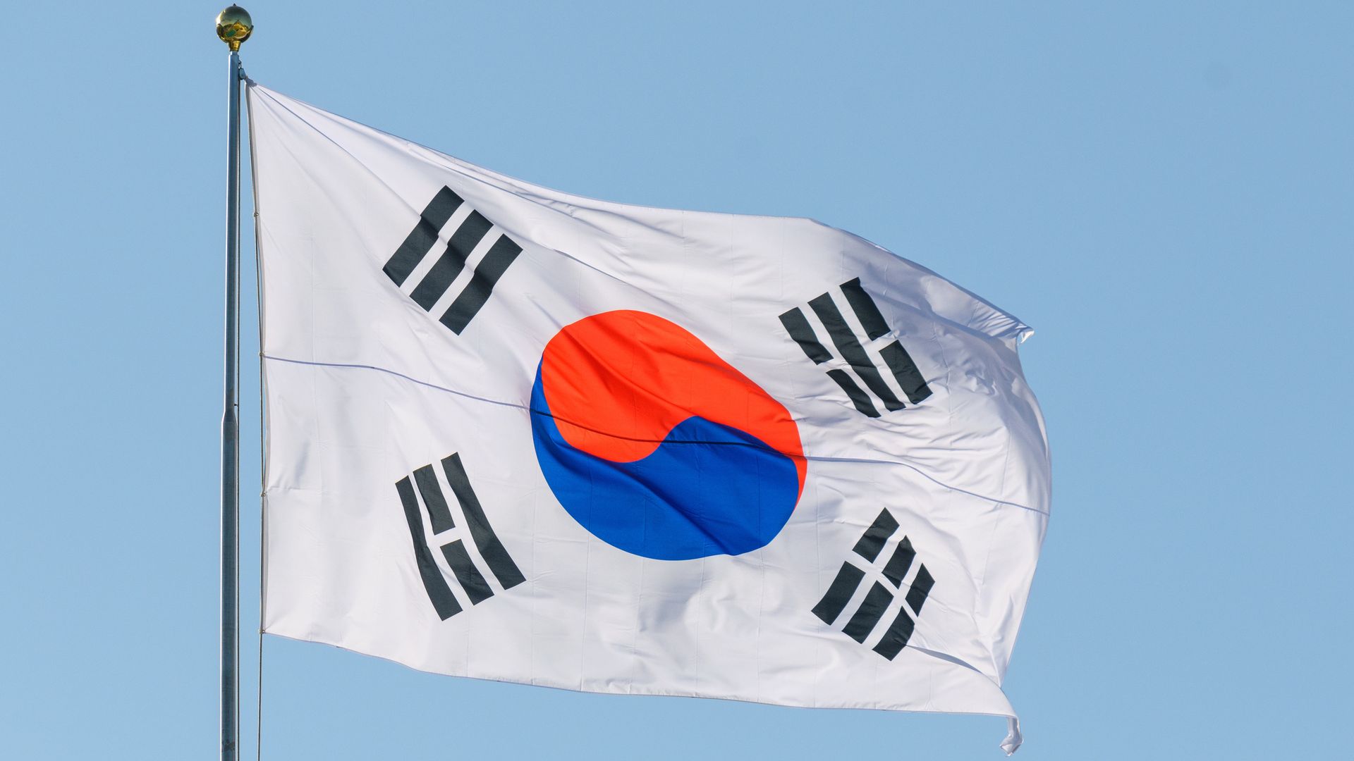 A South Korean flag, predominantly white in color, waves against a clear blue sky.