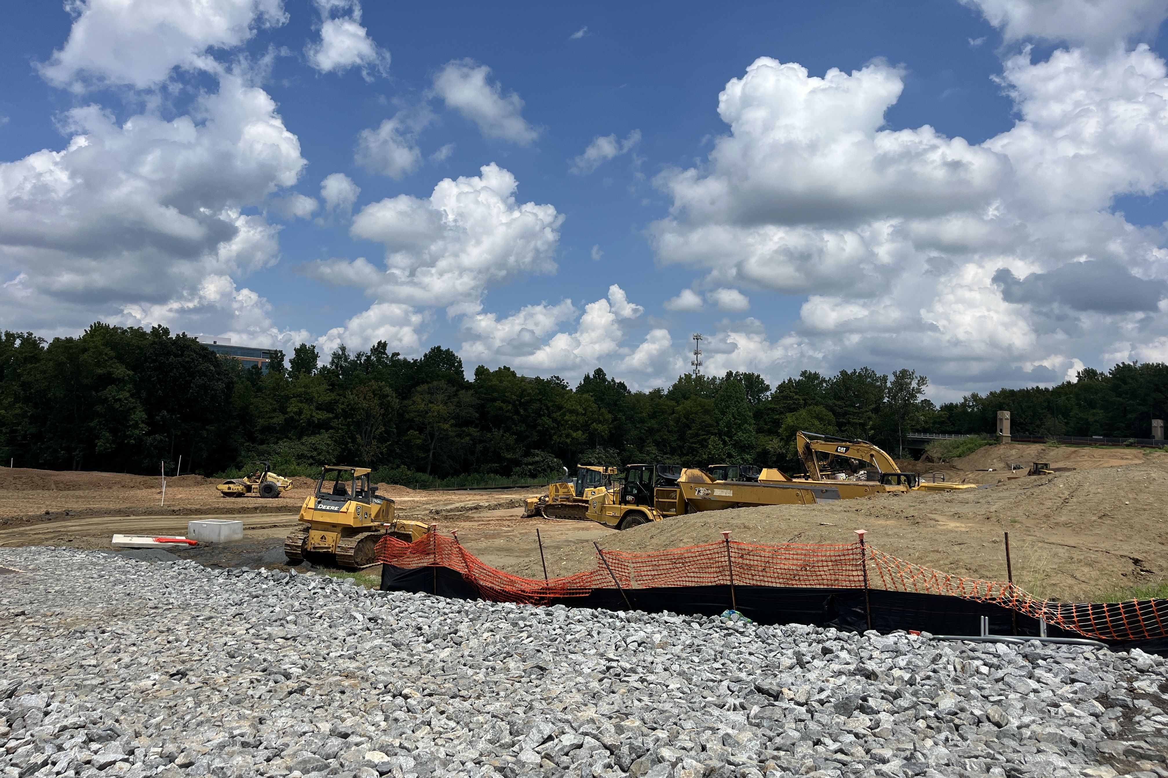 Construction site with yellow excavators and bulldozers on a dirt lot, orange safety fencing, and a gravel foreground. Tree line and blue sky with white clouds in the background.