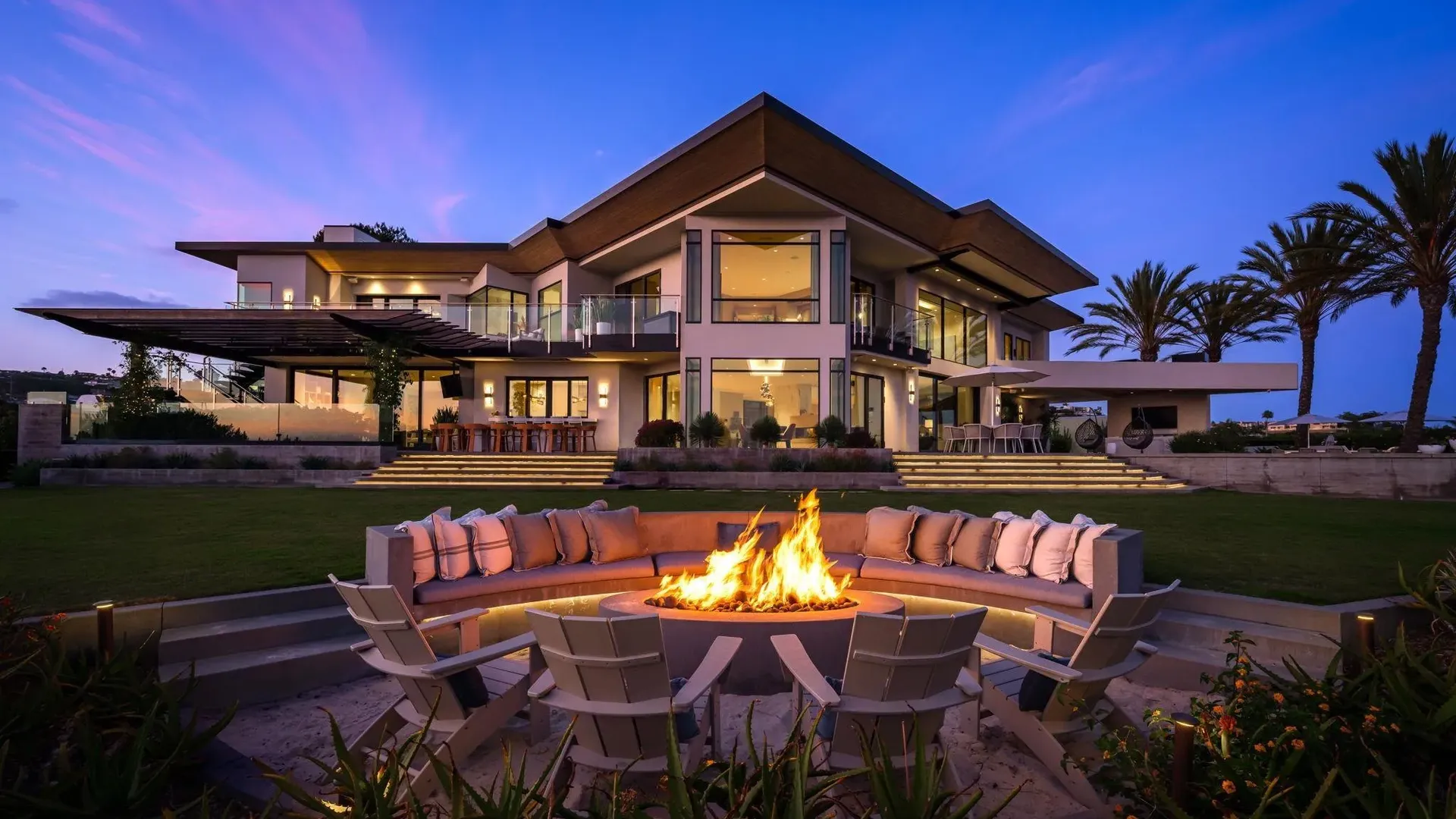 Modern two-story house with glass walls and balconies at dusk, a circular fire pit with orange flames surrounded by white chairs and couch, and palm trees on the right.