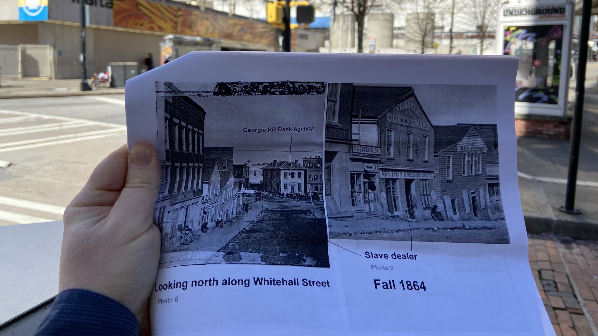 A hand holds a print out of photos from 1864 depicting a street scape in Atlanta with a store front offering to sell enslaved people