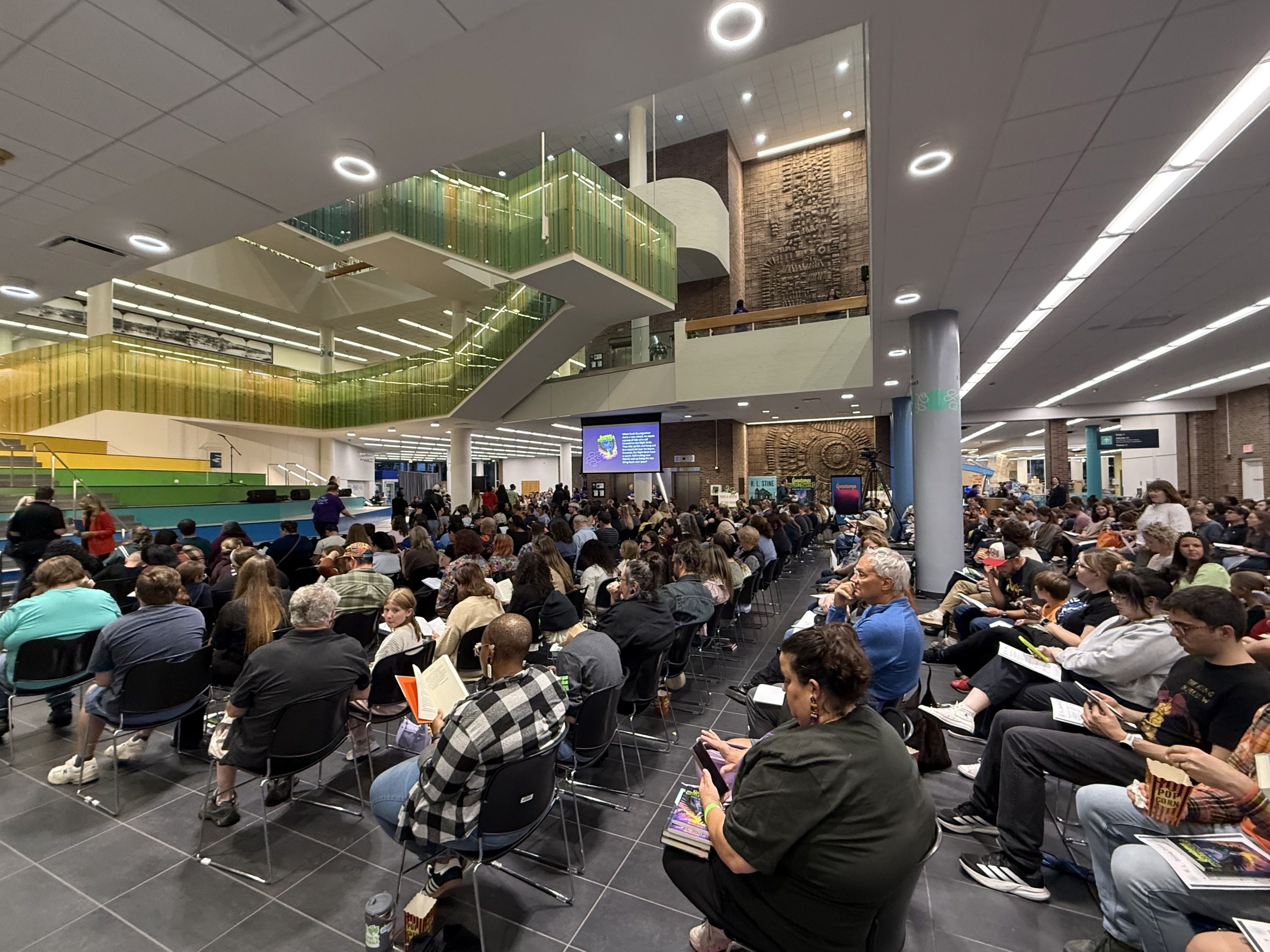 Crowded indoor event in a modern atrium: rows of chairs filled with people reading programs, a green glass balcony, brick walls, circular ceiling lights, and a distant stage.