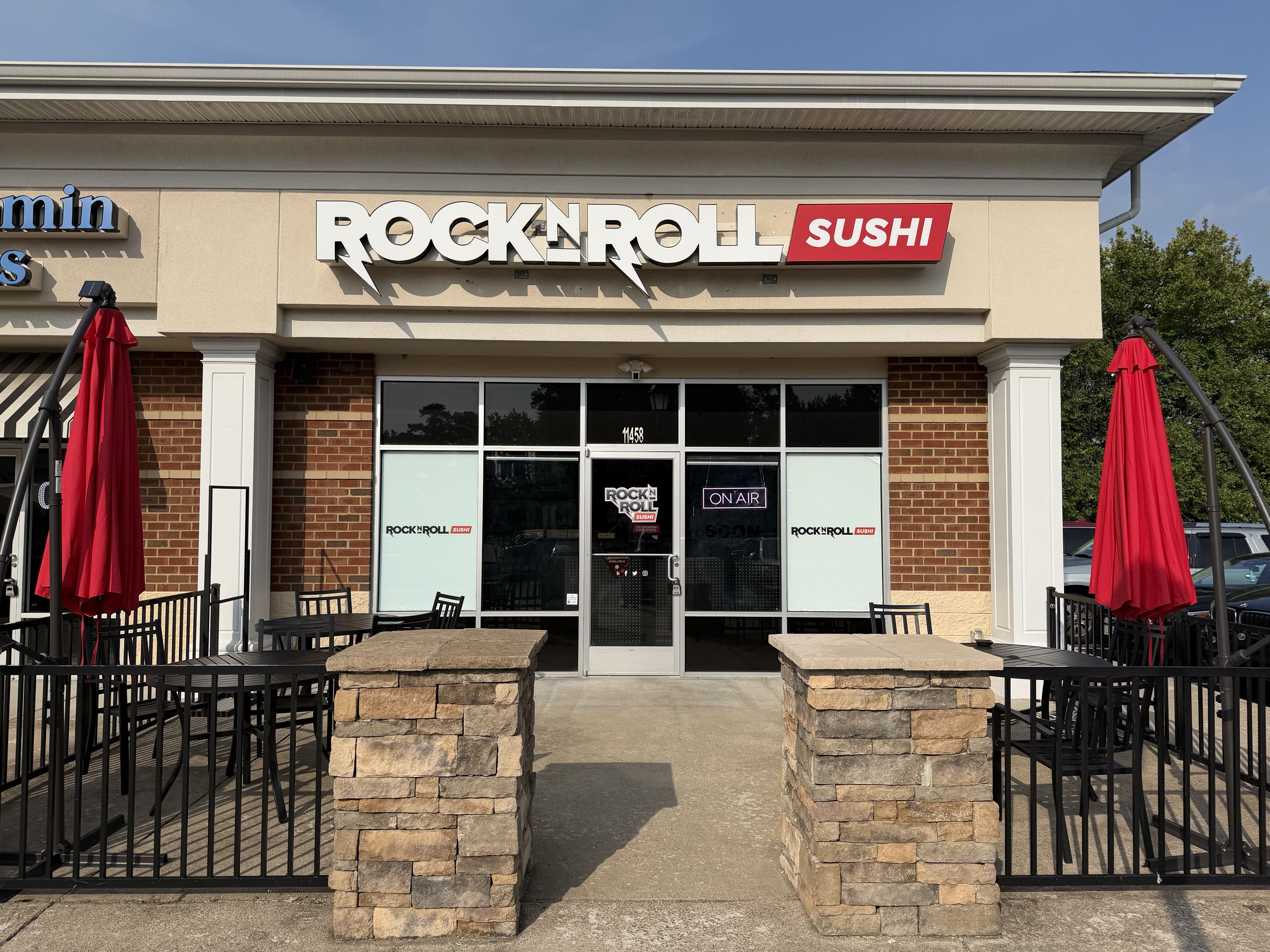 Exterior of Rock N Roll Sushi restaurant with a large white ROCK N ROLL sign and red SUSHI panel above glass doors; brick facade, outdoor seating with black tables and red umbrellas, blue sky.