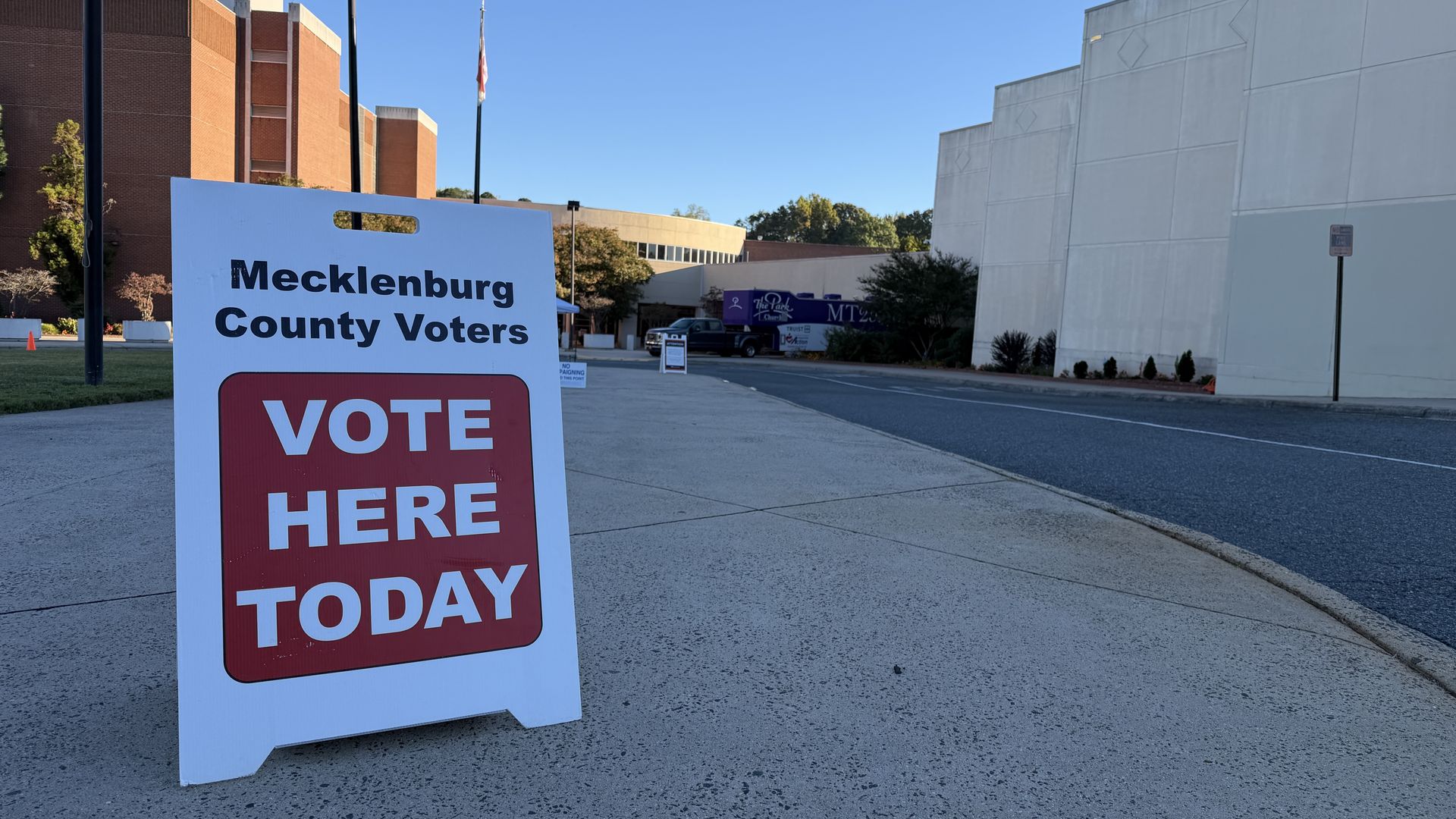 Foreground shows a white A-frame sign reading Mecklenburg County Voters with VOTE HERE TODAY in red; a street scene with brick buildings, a white wall, and a clear blue sky.