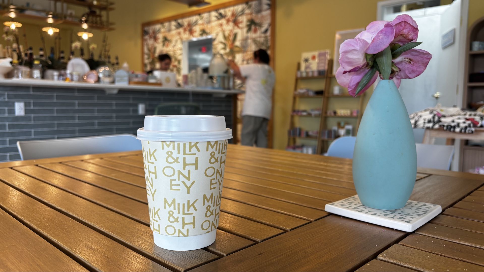 To-go coffee cup with a repeating "MILK & HONEY" design sits on a wooden table beside a light blue vase holding pink flowers; a busy cafe with yellow walls is blurred in the background.