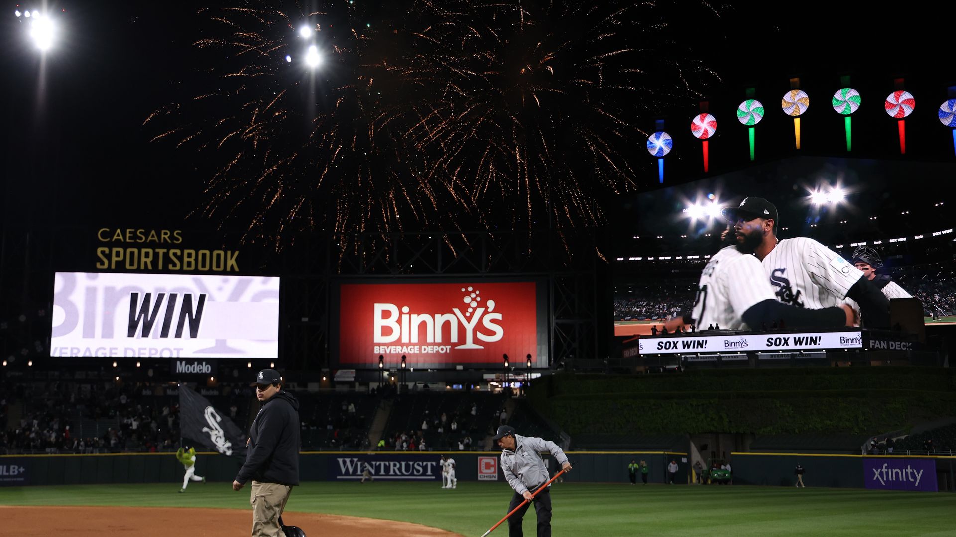 Night baseball game with fireworks over the stadium. Players on the field beneath bright floodlights; colorfully lit circular decorations on the stands and banners reading Sox Win and Binny's.